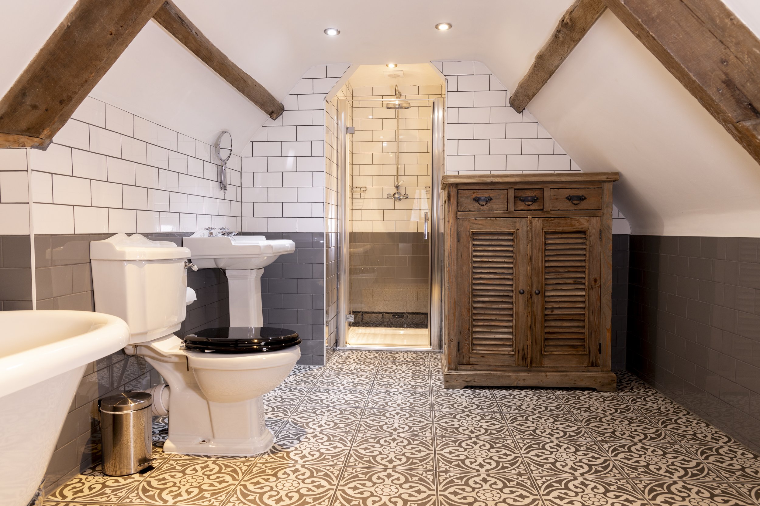 Rustic attic bathroom with exposed wooden beams, white subway tile walls, patterned floor tiles, a white toilet with a black seat, a pedestal sink, a glass shower enclosure, a wooden cabinet, and recessed lighting.