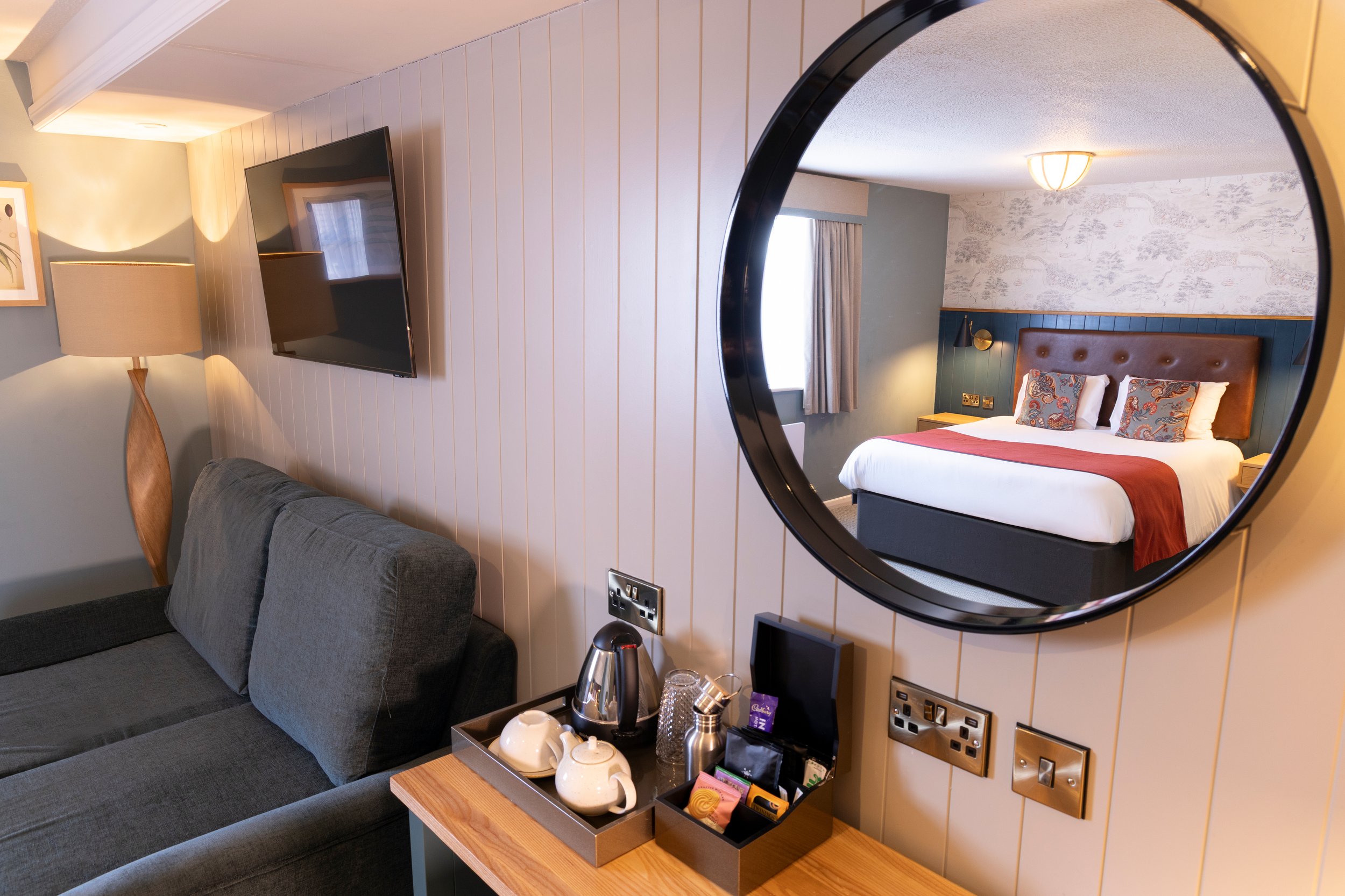 Modern hotel room with wall-mounted TV, blue sofa, floor lamp, and a reflection of a bed with decorative pillows in a round mirror. A wooden table with a tea set and kettle is also visible.