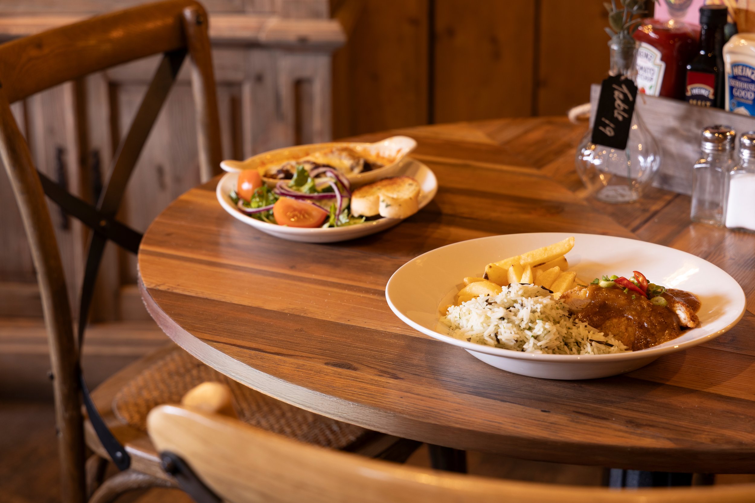 Wooden table with plates of food, including rice and chips with sauce, and salad with tomatoes and bread, inside a restaurant.