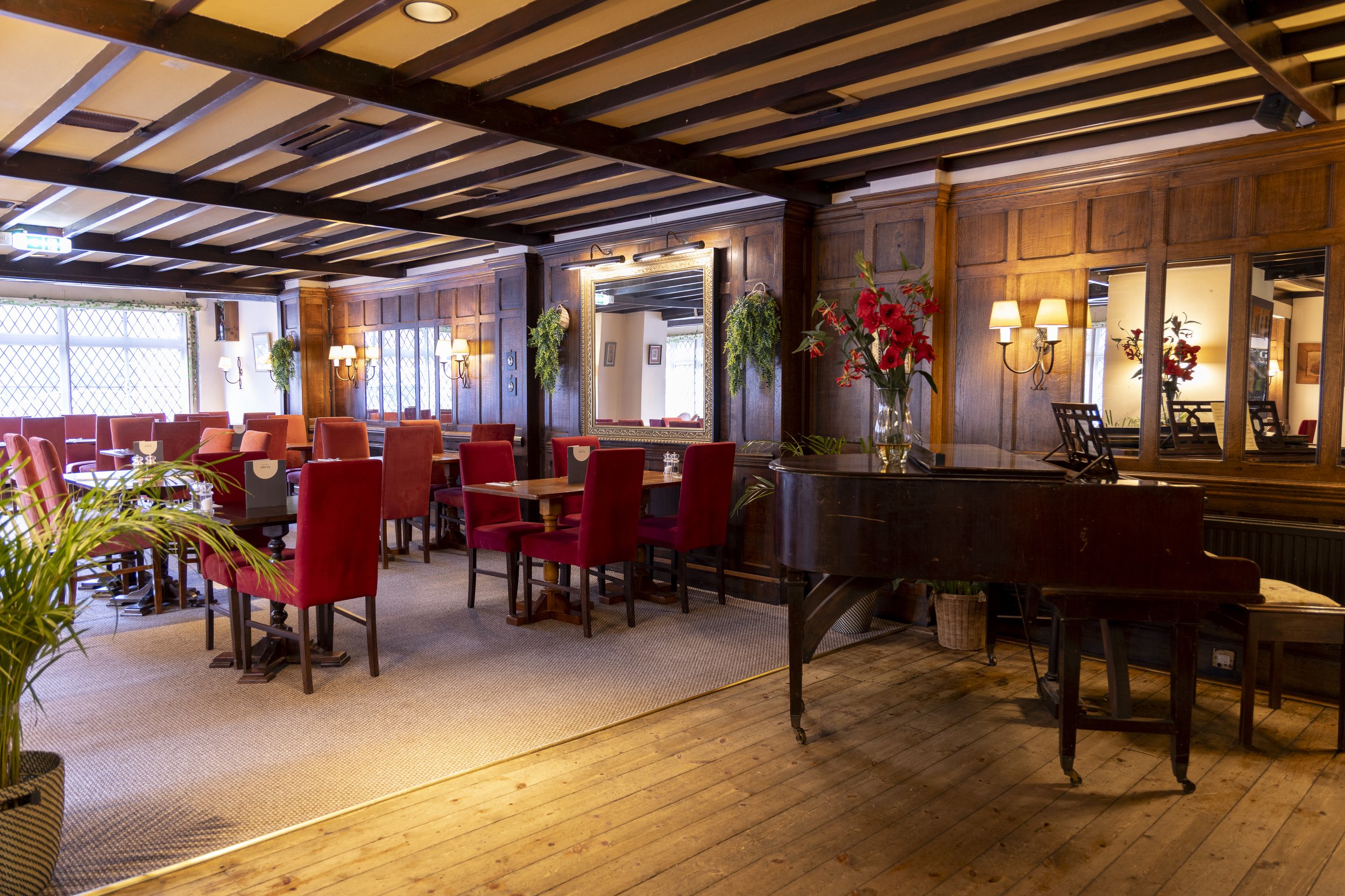 Interior of an elegant restaurant with wood-paneled walls, red upholstered chairs, and a grand piano with flowers on top. Mirrors and wall lamps adorn the walls, and houseplants add greenery to the decor.
