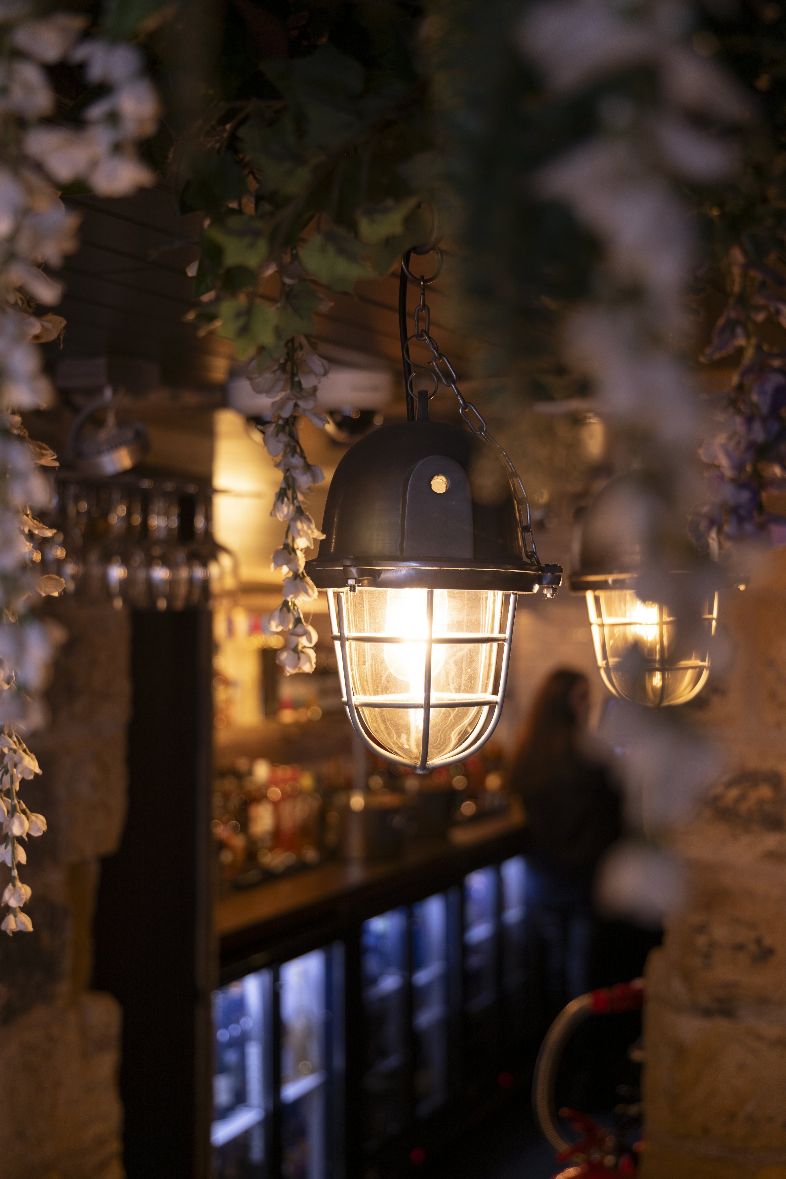 Vintage industrial-style pendant lights hanging in a dimly lit bar with exposed stone walls and shelves lined with liquor bottles.