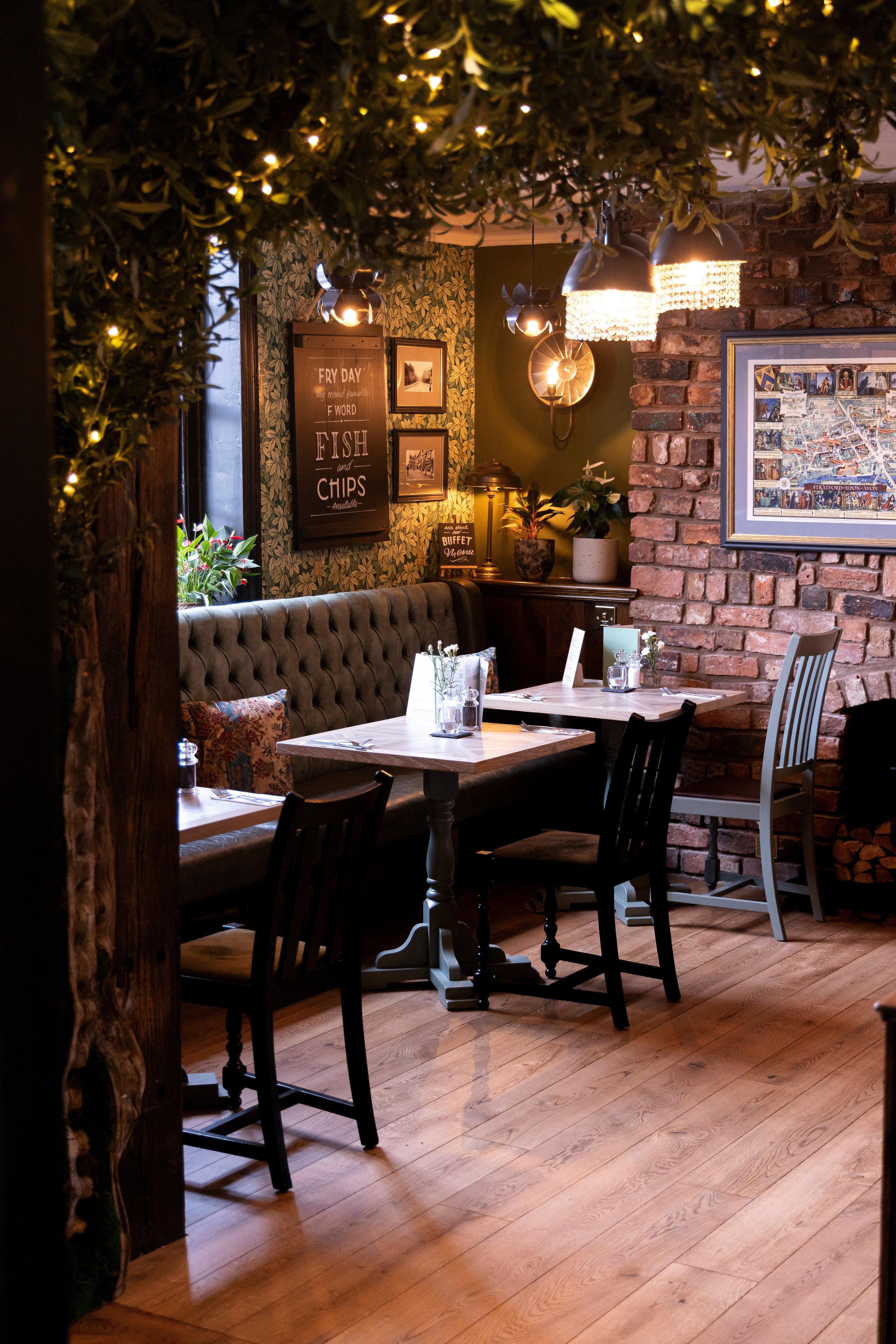 Cozy restaurant interior with wooden tables, chairs, and exposed brick walls, featuring a decorative wall with framed pictures and signs, potted plants, and ambient pendant lighting.