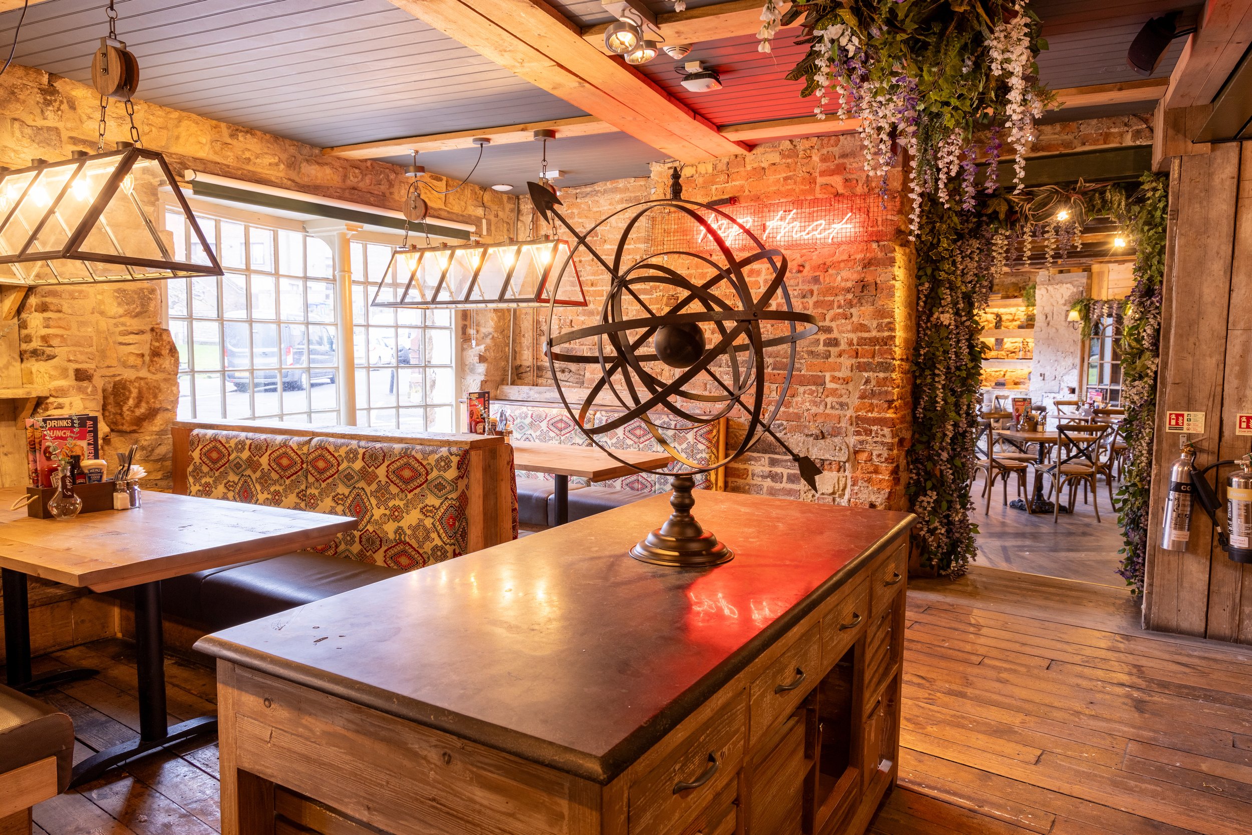 Cozy restaurant interior with rustic decor, wooden tables, patterned booth seating, vintage lights, decorative globe sculpture, brick walls, greenery, and a neon sign.