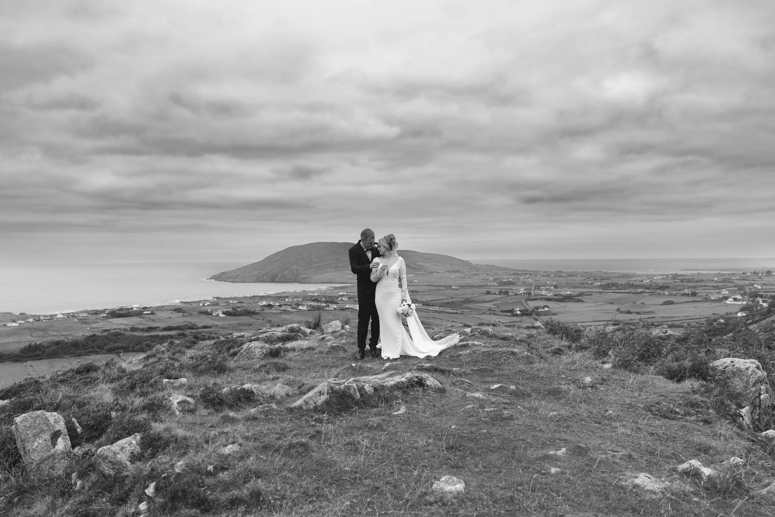 Black and white photo of a bride and groom standing on a hill with a scenic landscape, ocean, and mountains in the background. The groom is dressed in a dark suit, and the bride in a white wedding dress holding a bouquet.