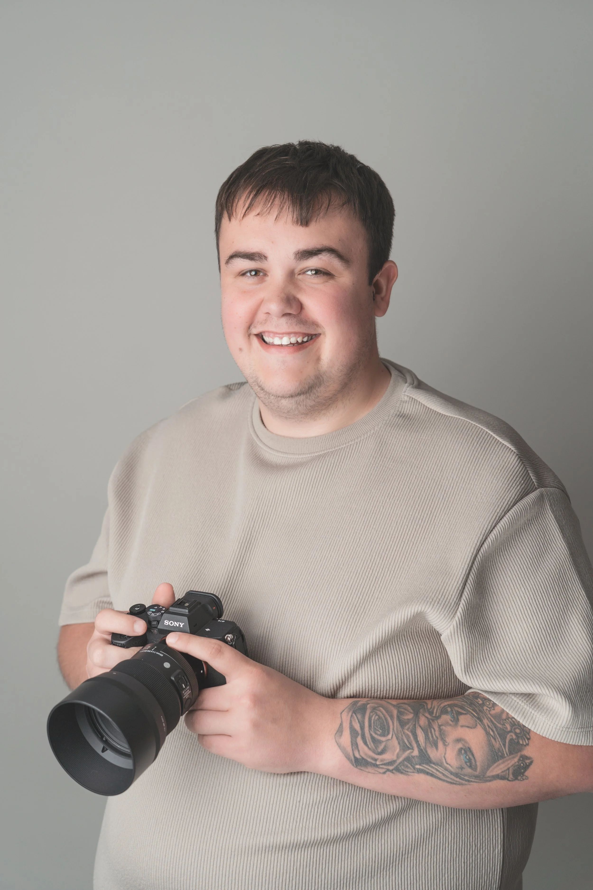 A young man smiling, holding a professional camera, with a tattoo sleeve on his left arm, wearing a beige shirt, standing against a plain gray background.