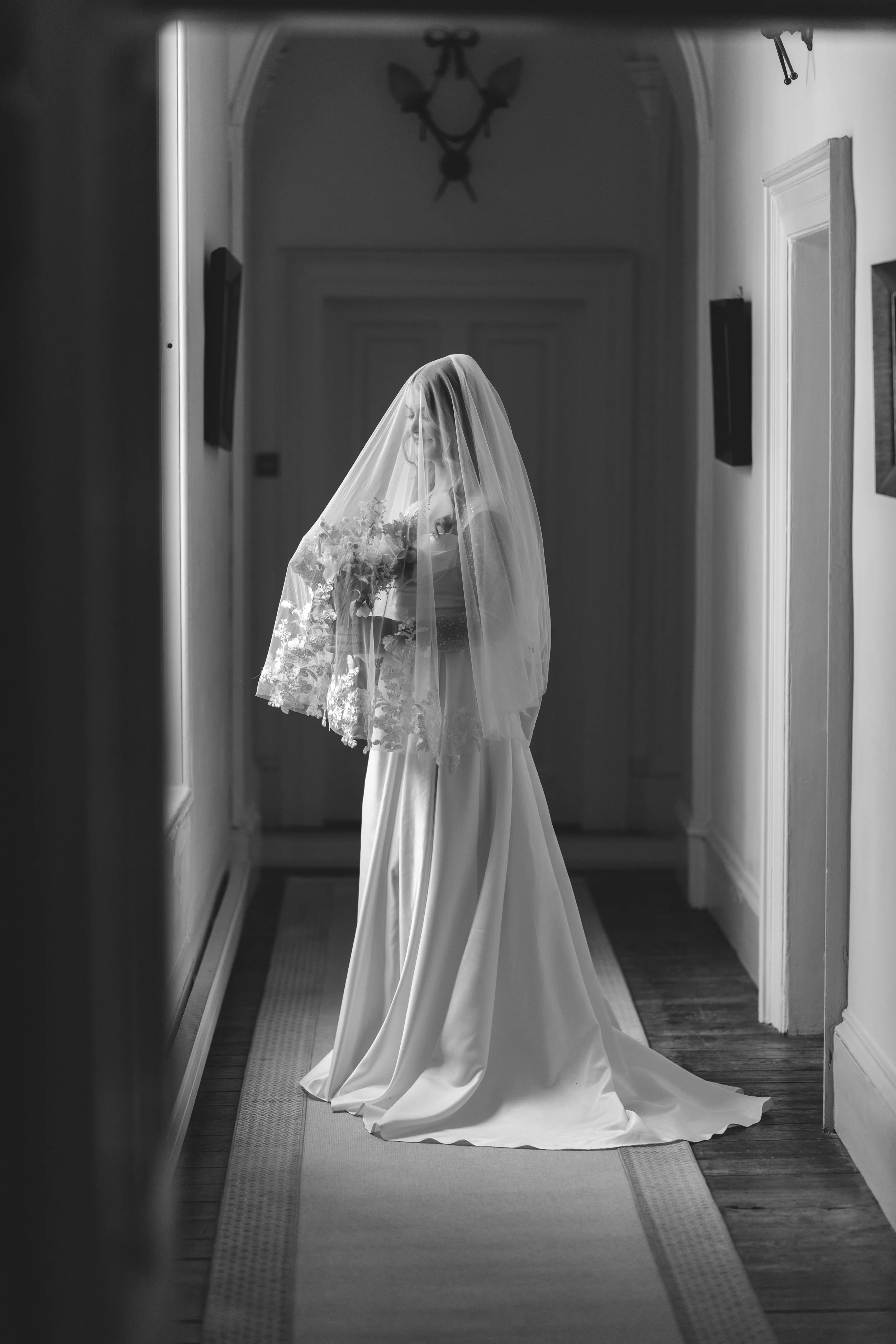 Black and white photo of a bride in a wedding dress with a veil, holding a bouquet, standing in a hallway.