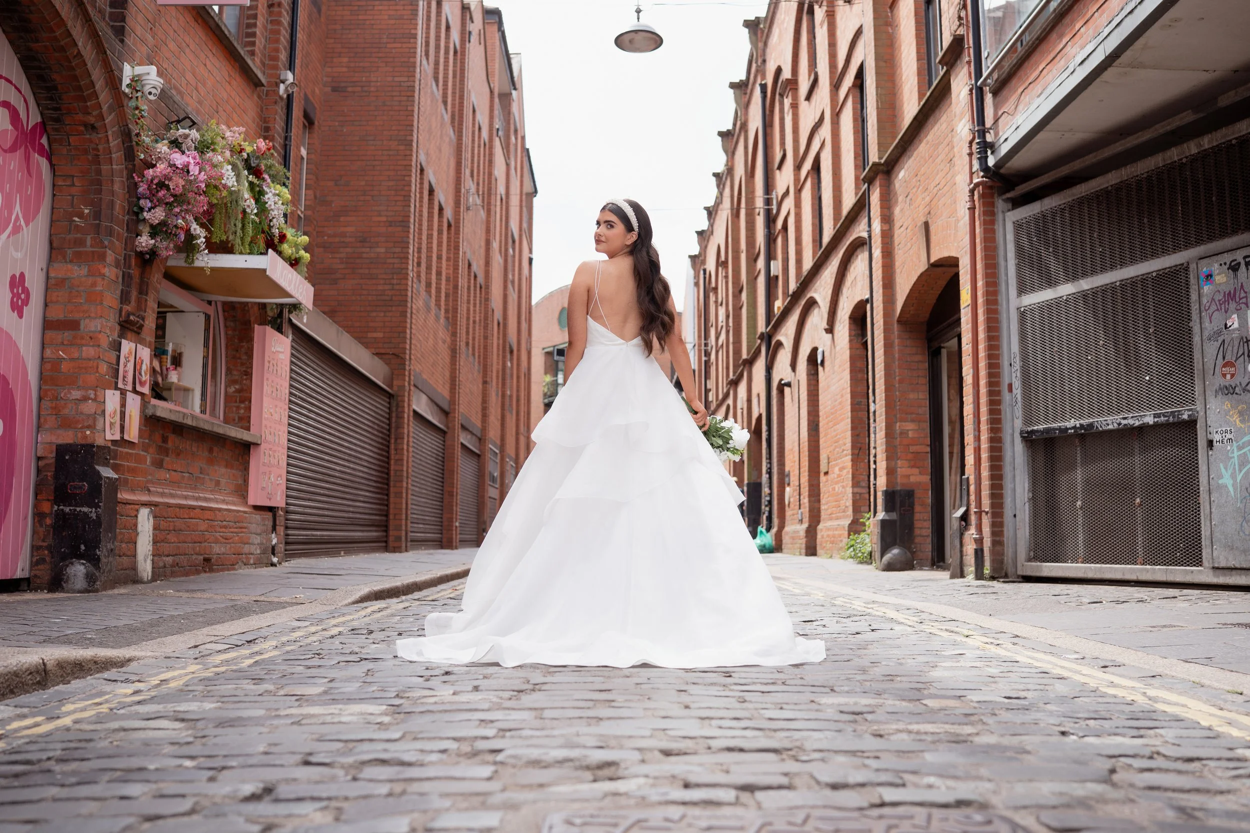 A woman wearing a white wedding dress walking on a cobblestone street in an urban area with brick buildings. She is holding a bouquet of flowers and looking back over her shoulder.