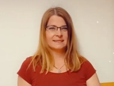 Das Bild zeigt eine Frau mit rotblonden, schulterlangen Haaren und rotem T-shirt vor einem weißen Hintergrund. Sie trägt eine Brille. 