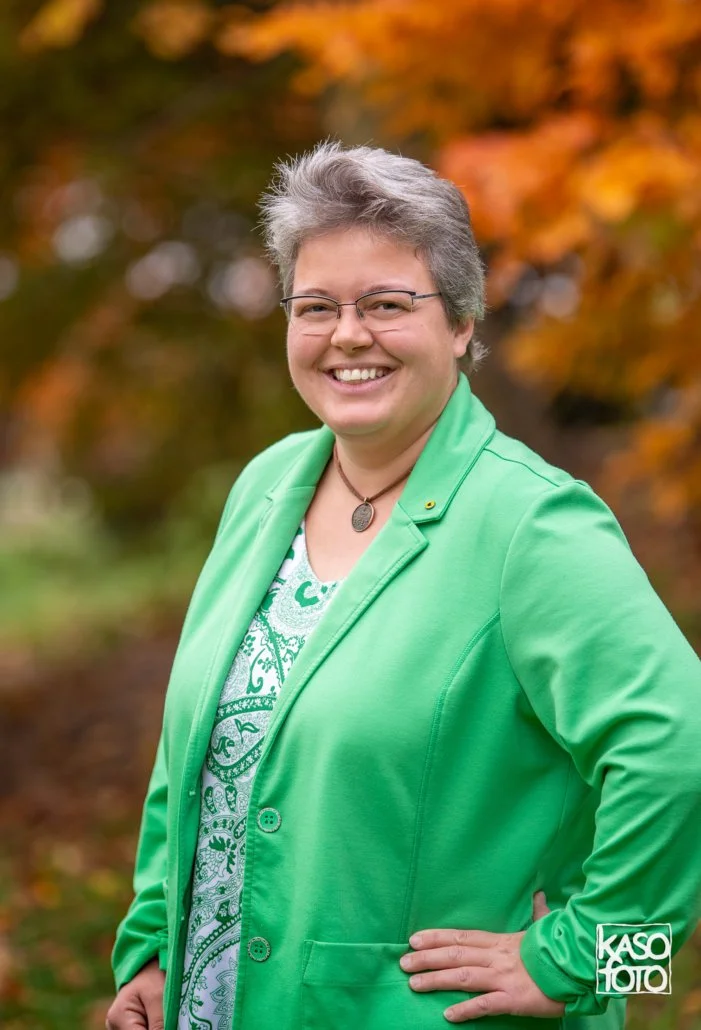 Das Bild zeigt eine freundlich lächelnde Frau mit grünweißen T-Shirt und einem Grünen Blazer und grauen kurzen Haaren und einer Brille.  Verschwommen im Hintergrund ist ein Baum mit orangeroten Blättern.