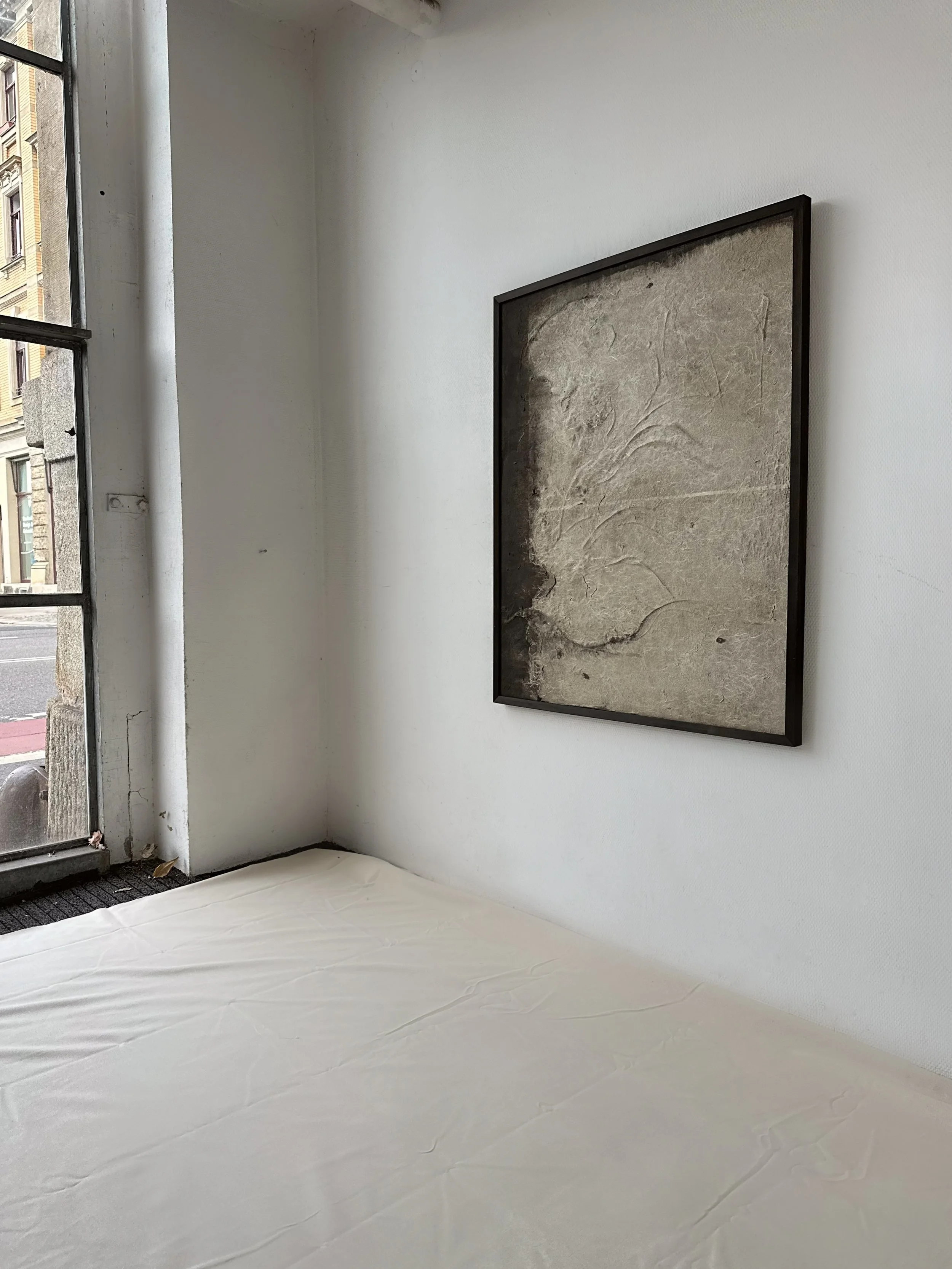 Ein weißes Bett mit glatter Bettwäsche, eine weiße Wand mit einem großen, abstrakten Gemälde in einem schwarzen Rahmen, neben einem großen Fenster mit Blick auf eine Straße und altbäckeren Gebäuden.