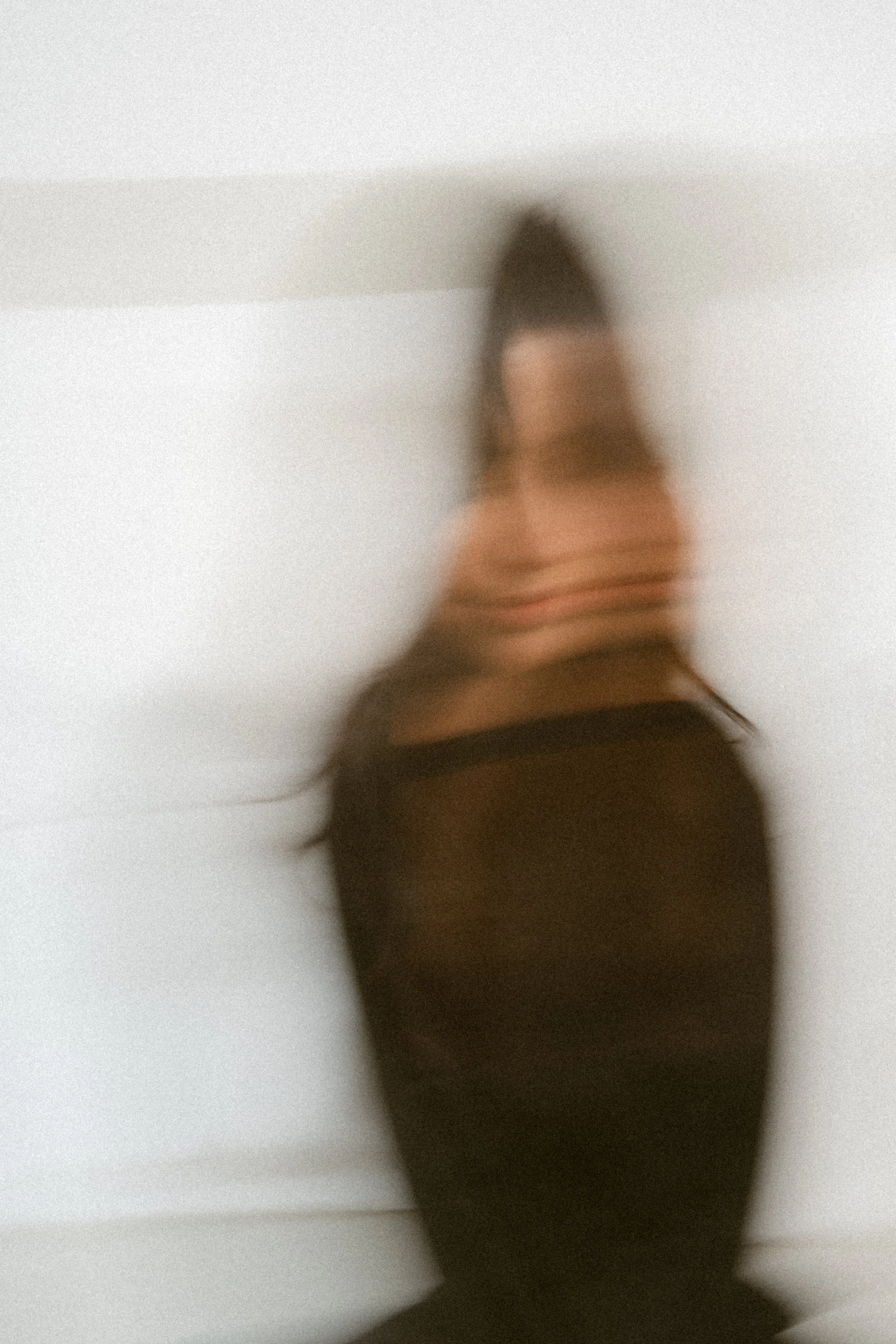 Ein unscharfes Foto einer Frau, die vor einem weißen Hintergrund steht, mit langen dunklen Haaren und dunkler Kleidung.