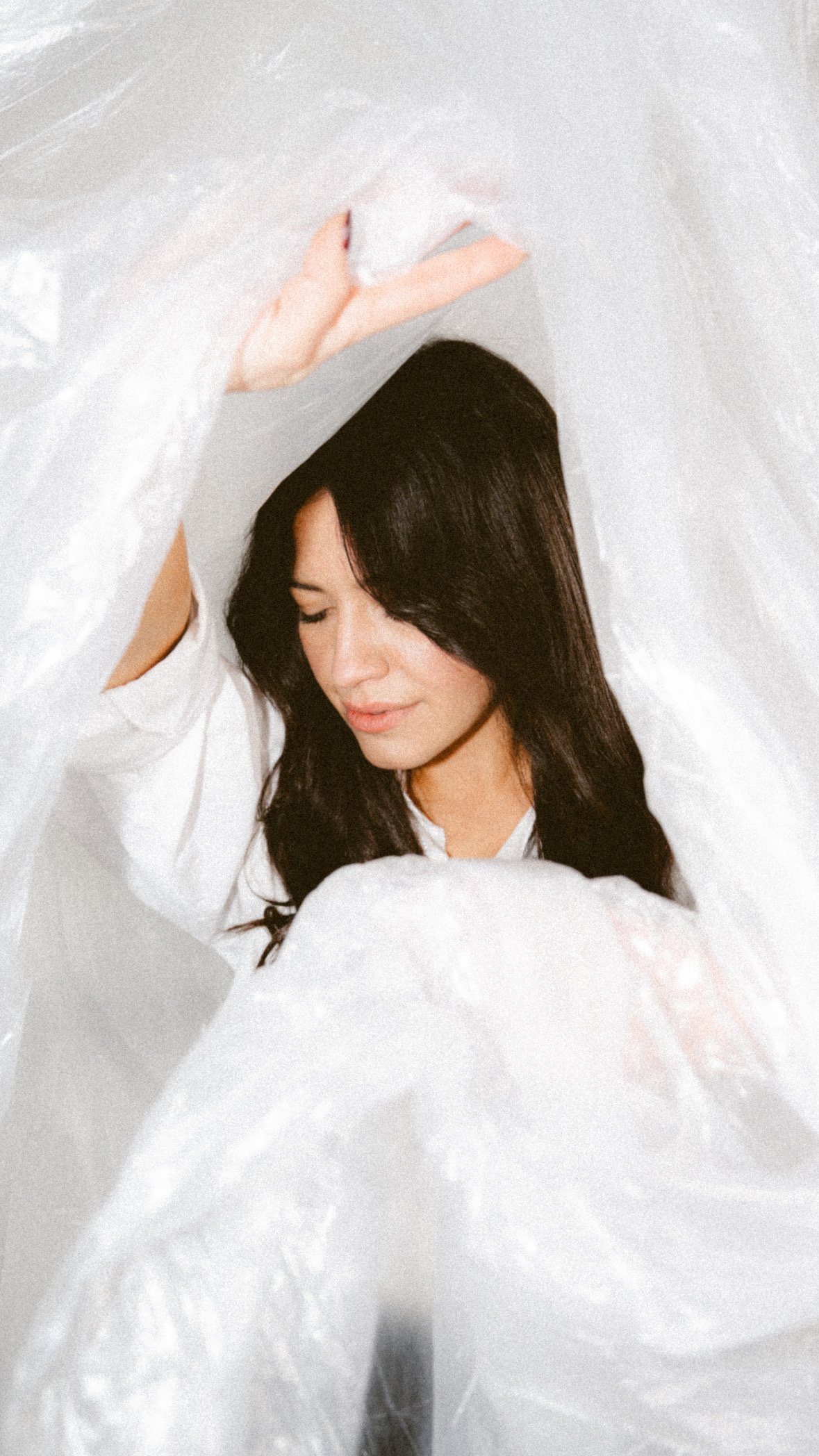 Frau mit dunklen Haaren, die in einem weißen Kleid unter einer weißen, durchsichtigen Decke steht oder sitzt.