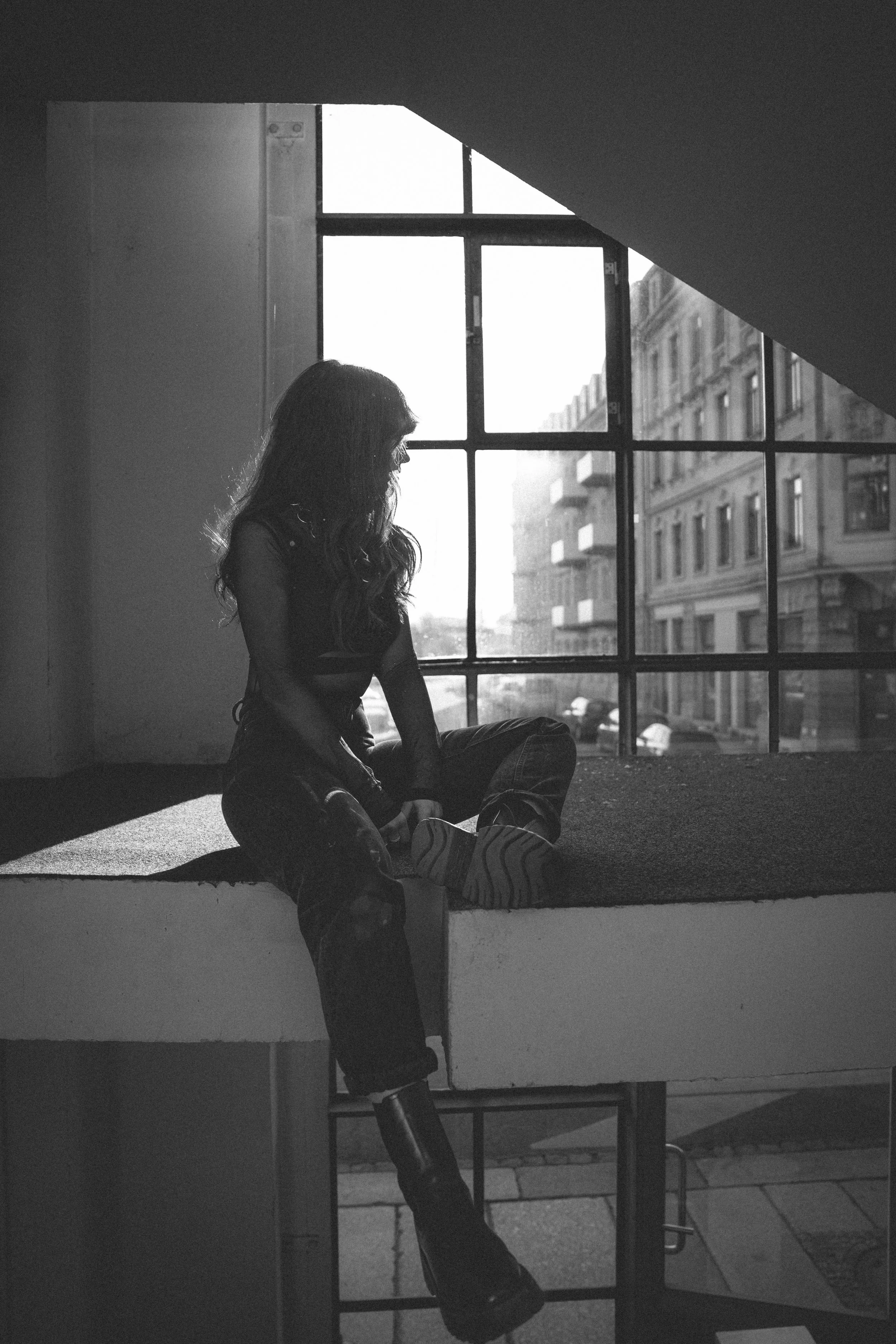 Schwarze und weiße Aufnahme einer jungen Frau in Ihrem Atelier, die auf einem Fensterbrett sitzt, während die Sonne draußen scheint, Blick nach links, urbaner Hintergrund.