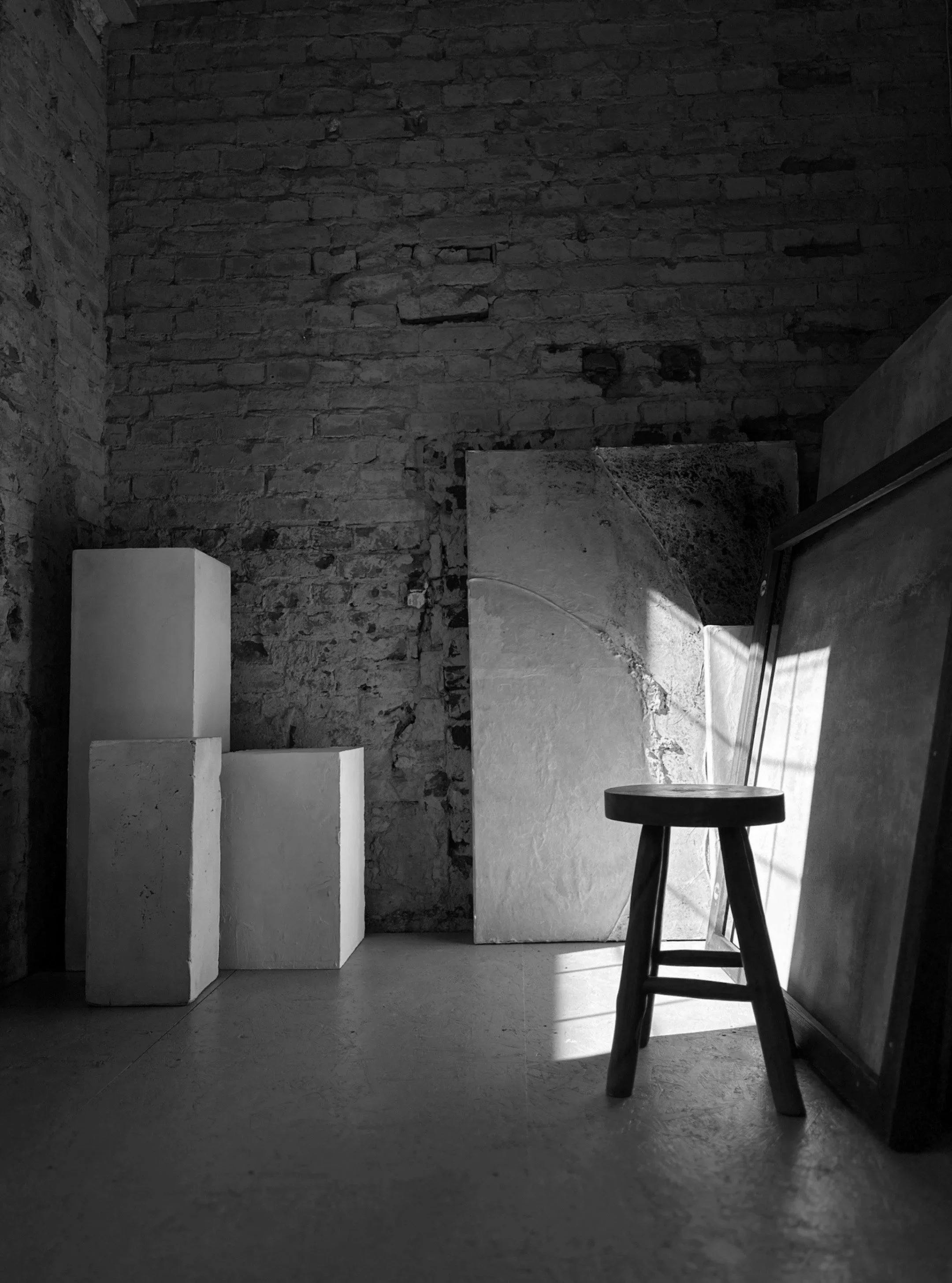 Schwarz-weiß-Foto eines Raumes mit Feldsteinmauer, leeren Skulpturen, einem Hocker und mehreren Leinwänden, die an die Wand gestellt sind.