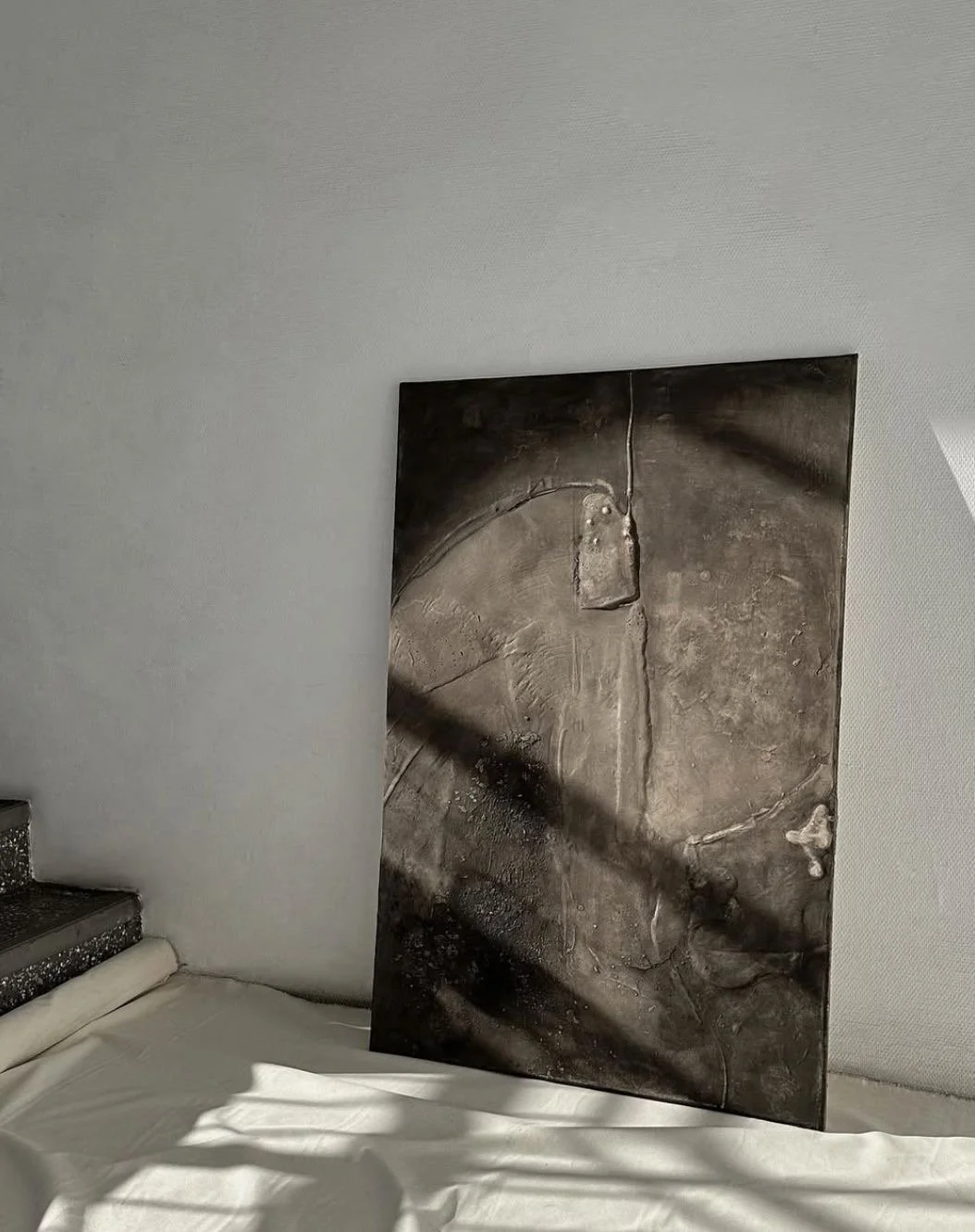 Ein dunkles abstraktes Gemälde steht auf einem weißen Bett, neben einer Treppe in einem hellen Raum.