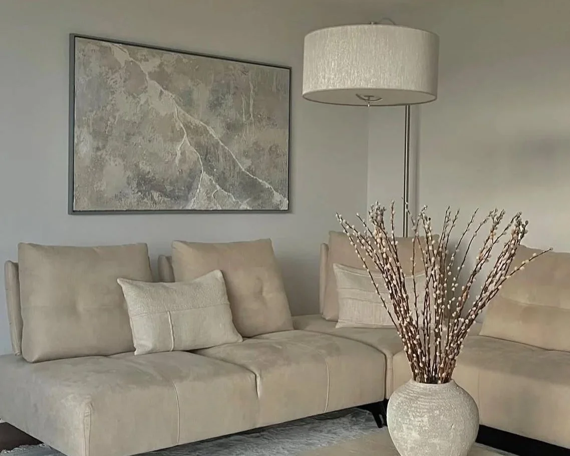 Ein Wohnzimmer mit einem beige Sofa, einer Vase mit Zweigen, einer Stehlampe und einem gemalten Bild an der Wand.