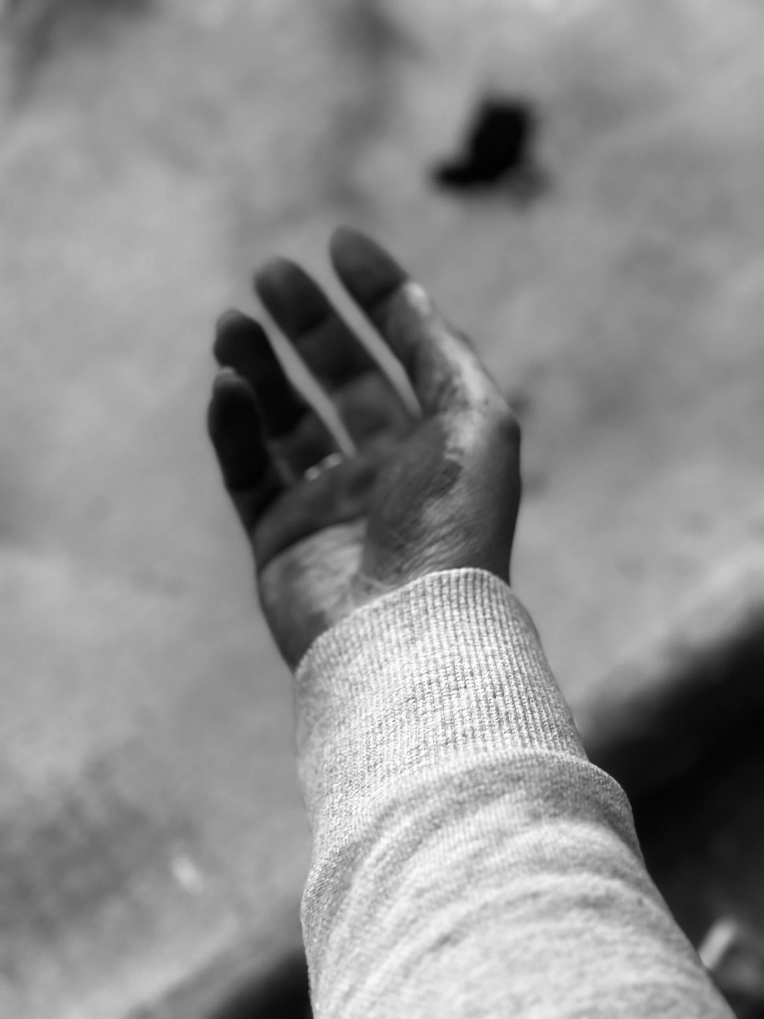 Schwarz-weiß-Foto eines ausgestreckten Hand mit einem Ring am Finger, das auf den Boden zeigt, im Hintergrund eine schwarze Socke auf einem Bodenbelag.