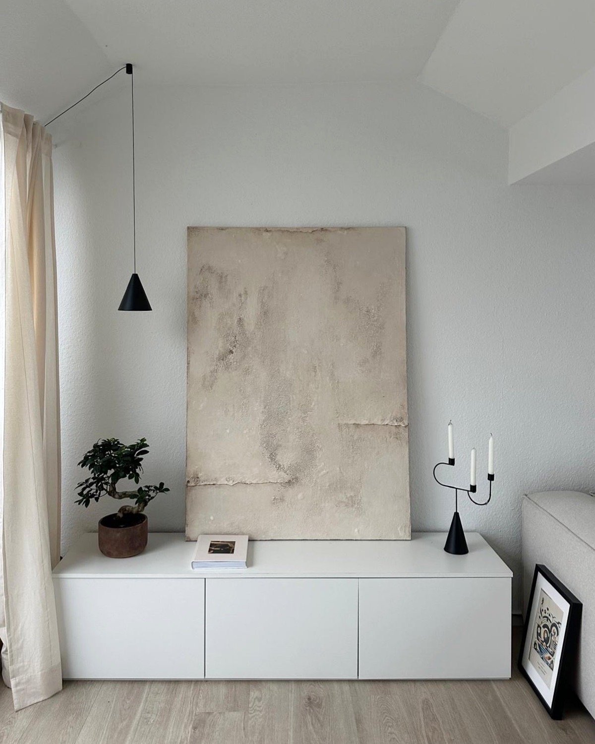 Ein Wohnzimmer mit weißem Sideboard, einer kleinen Bonsai-Pflanze, einem Buch, einem schwarzen Kronleuchter und einem großen, abstrakten Wandbild. Es gibt auch eine stehende Lampe und ein gerahmtes Bild auf dem Boden.