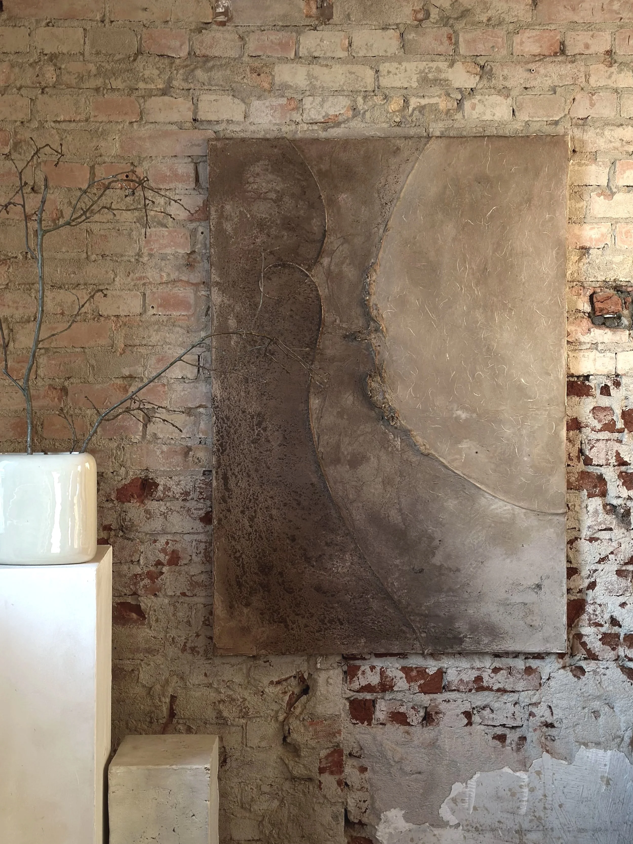 Abstraktes Wandbild mit erdigen Tönen, hängt an einer Backsteinwand, neben einer weißen Vase mit dünnen, trockenen Zweigen.