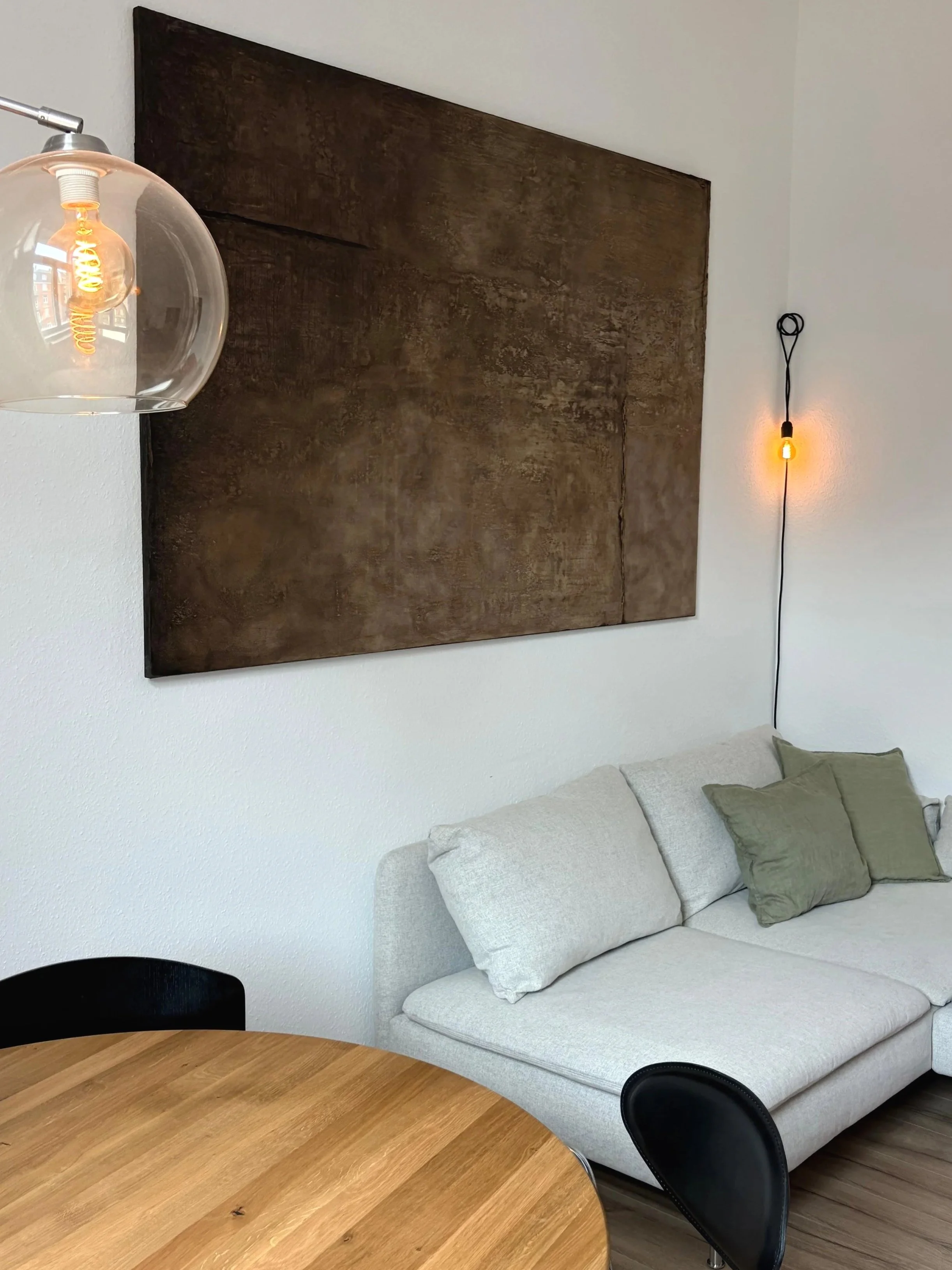 Ein modern eingerichteter Raum mit einem weißen Sofa, grünen Kissen, einem runden Holztisch und einer dunkelbraunen Wanddeko. Es gibt eine warme Wandlampe und eine Glühlampe in einer Glasleuchte.