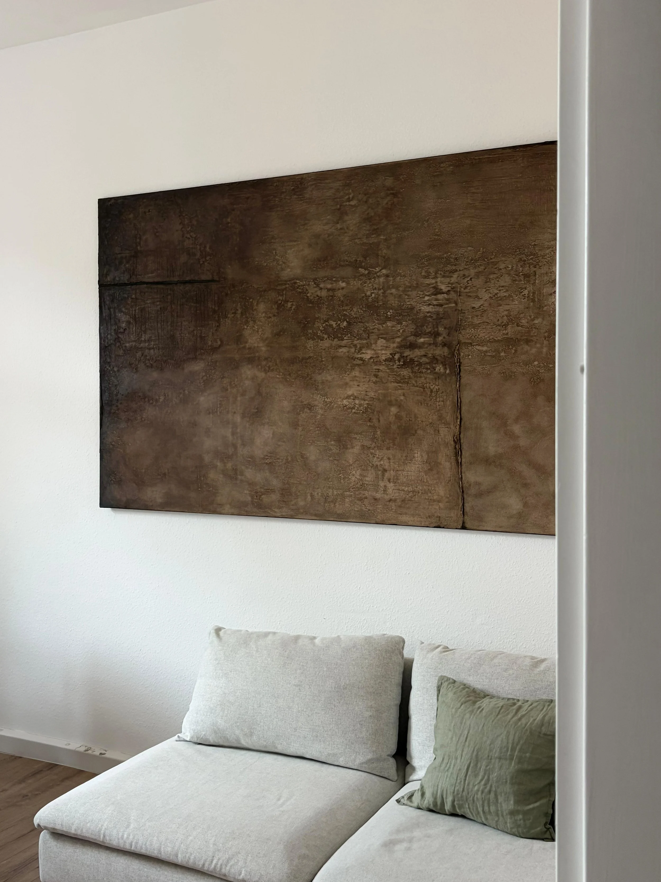 Ein Wohnzimmer mit weißer Wand, einem großen rechteckigen Holzbild an der Wand und einem weißen Sofa mit zwei Kissen, eines grau und eines grün.