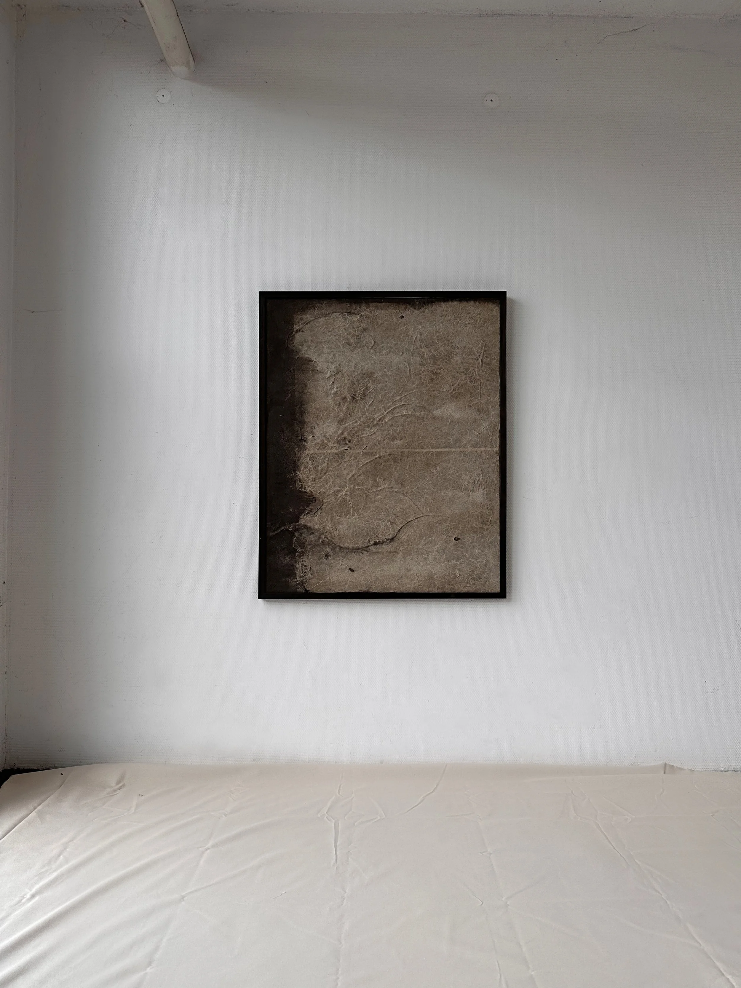 Ein leeres, weißes Zimmer mit einem schwarzen Rahmen an der Wand und einem weißen Bett. In der Mitte der Wand hängt ein abstraktes Kunstwerk, das wie eine vernarbte oder verwitterte Oberfläche aussieht.