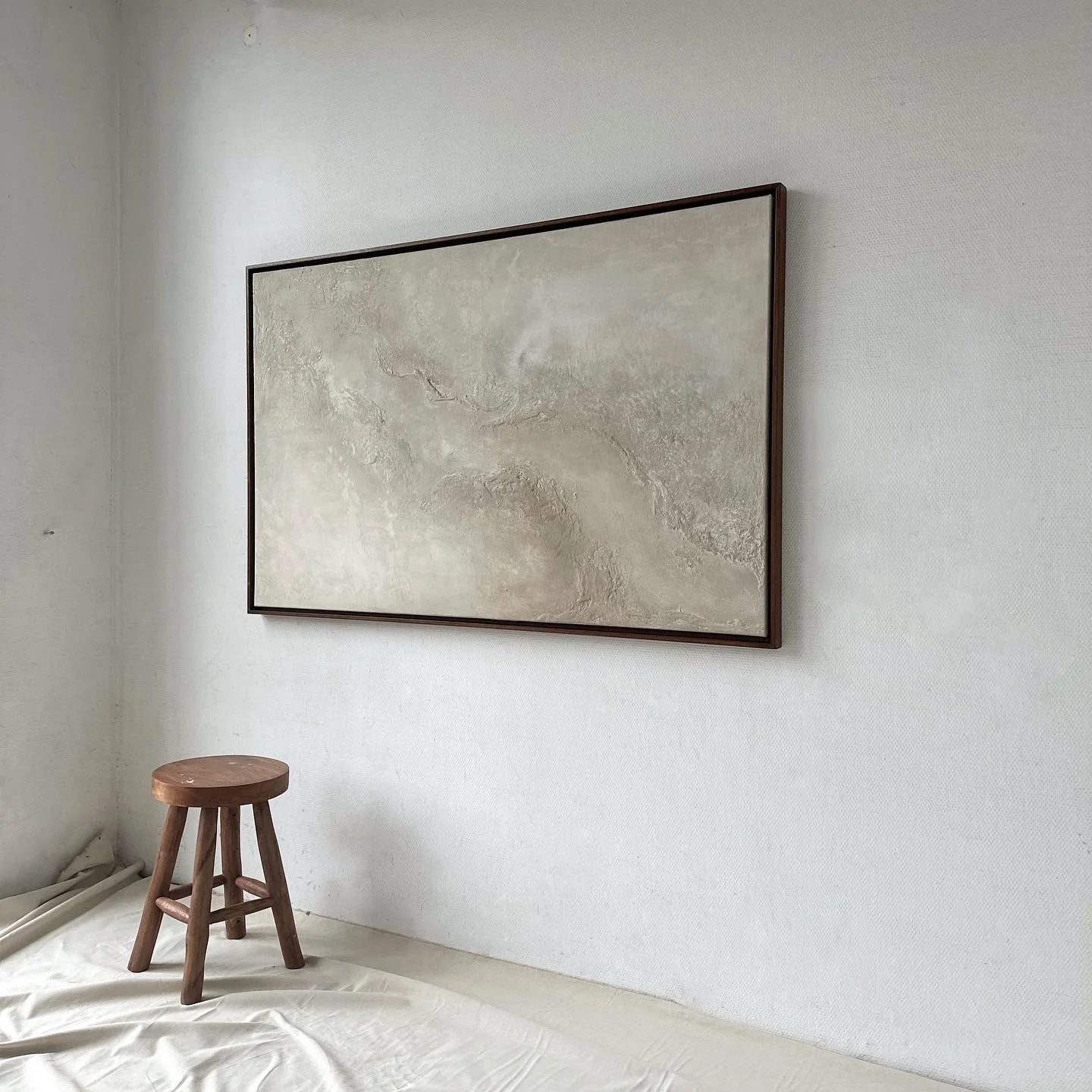 Ein leeres Bild in einem Holzrahmen an einer weißen Wand, daneben ein kleiner Holzschemel, auf dem Boden liegt eine weiße Decke.