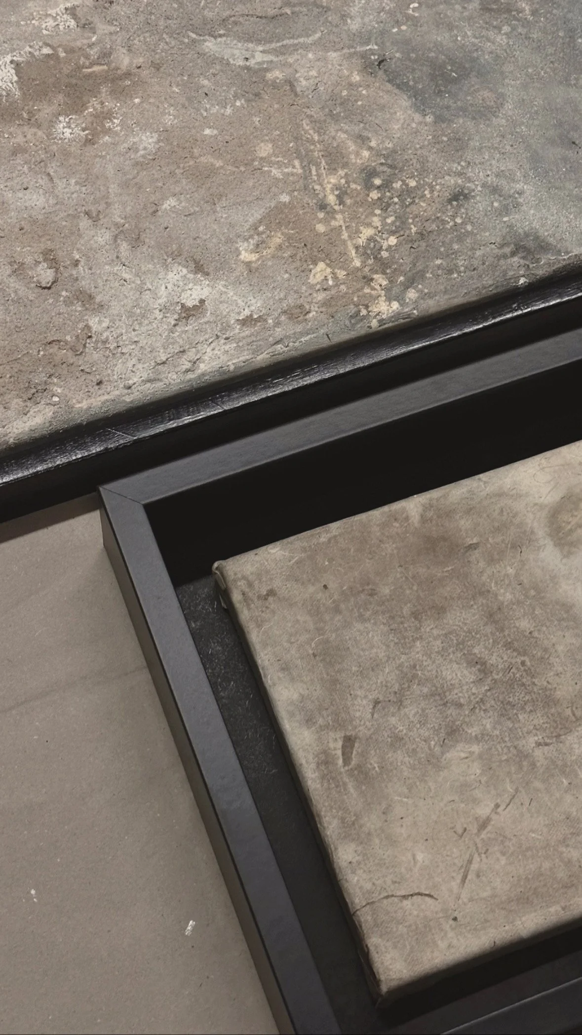 Ein schwarzer Rahmen mit einem grauen, schmutzigen Kissen oder Polster darauf, daneben ein Bodenbelag mit Grautönen und einer rauen Textur.