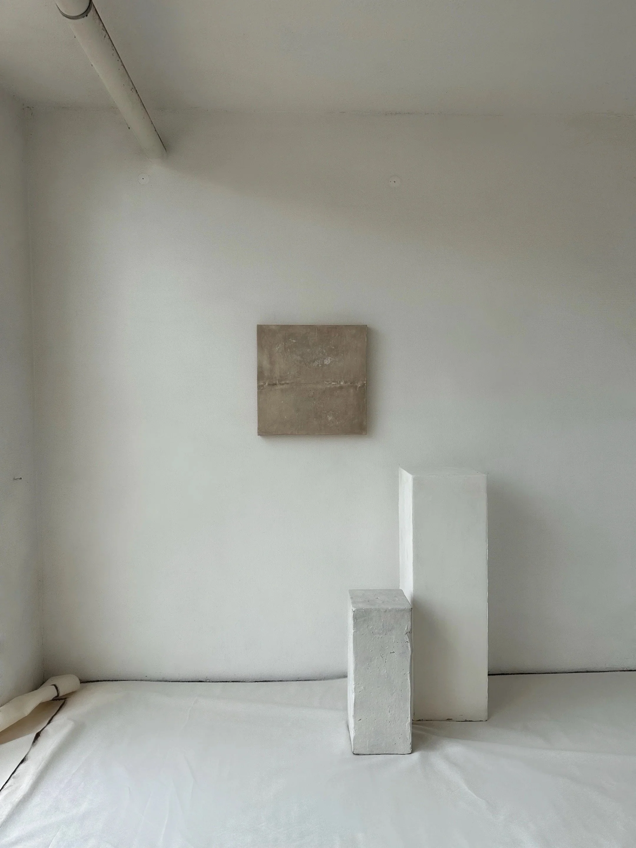 Ein minimalistischer Raum mit weißen Wänden und Boden, auf dem drei unterschiedlich hohe rechteckige Blöcke aus Stein oder Beton stehen, und eine kleine quadratische Platte an der Wand.