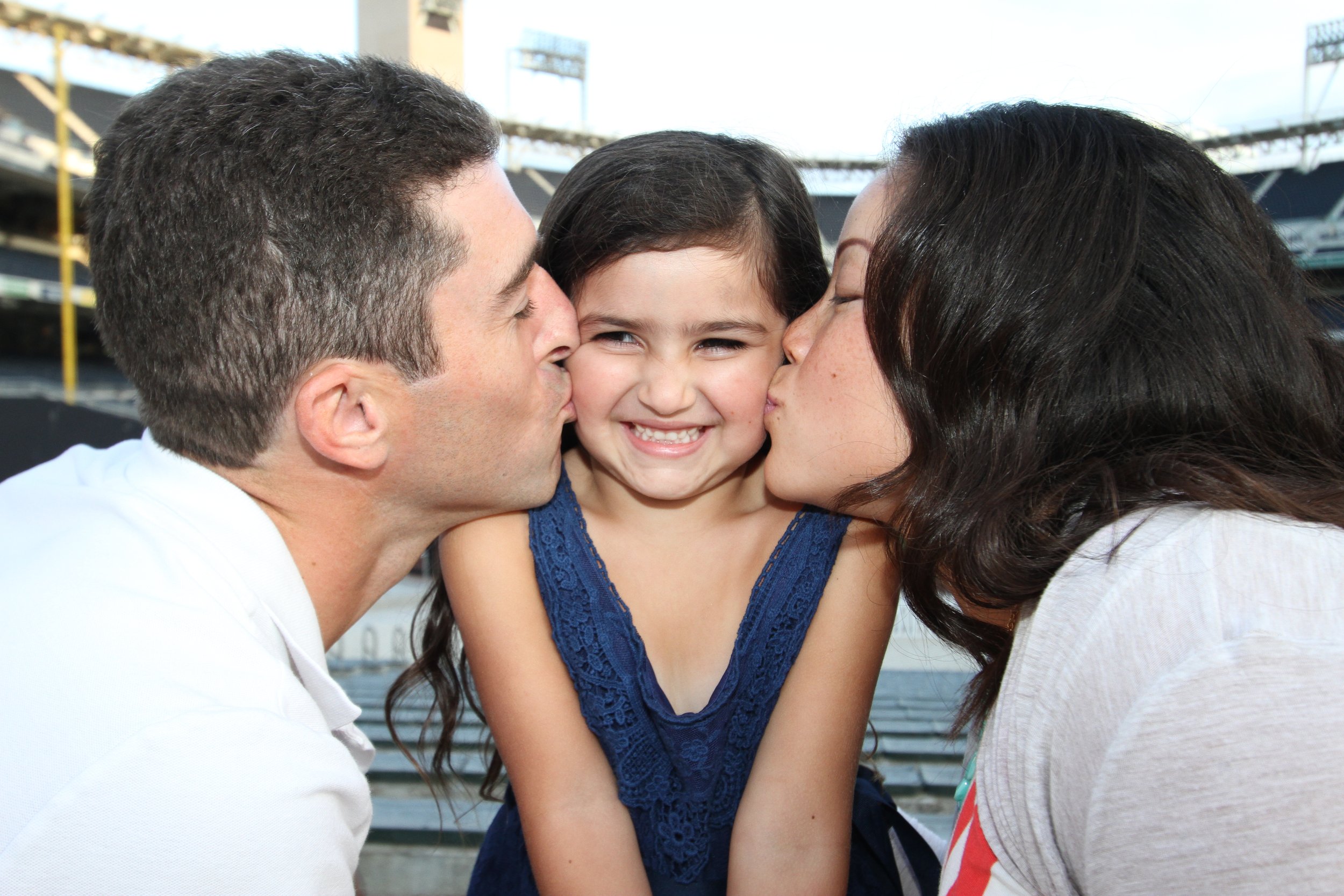 Family photoshoot capturing parents kissing their daughter on the cheeks in front of Petco Park in San Diego, celebrating family bonds.