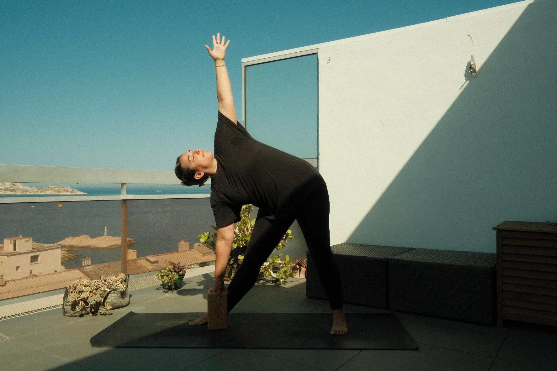 Femme faisant une posture de yoga sur un balcon avec vue sur la mer, portant un t-shirt noir et des leggings noirs, utilisant un bloc pour soutenir sa main, sous un ciel bleu clair.