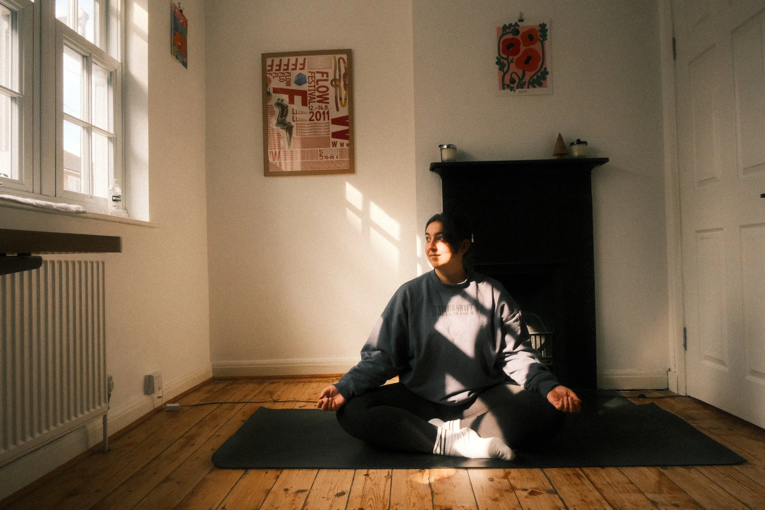 Femme assise en position de méditation sur un tapis de yoga dans une pièce lumineuse avec fenêtres, murs blancs, décorations murales colorées et cheminée noire.