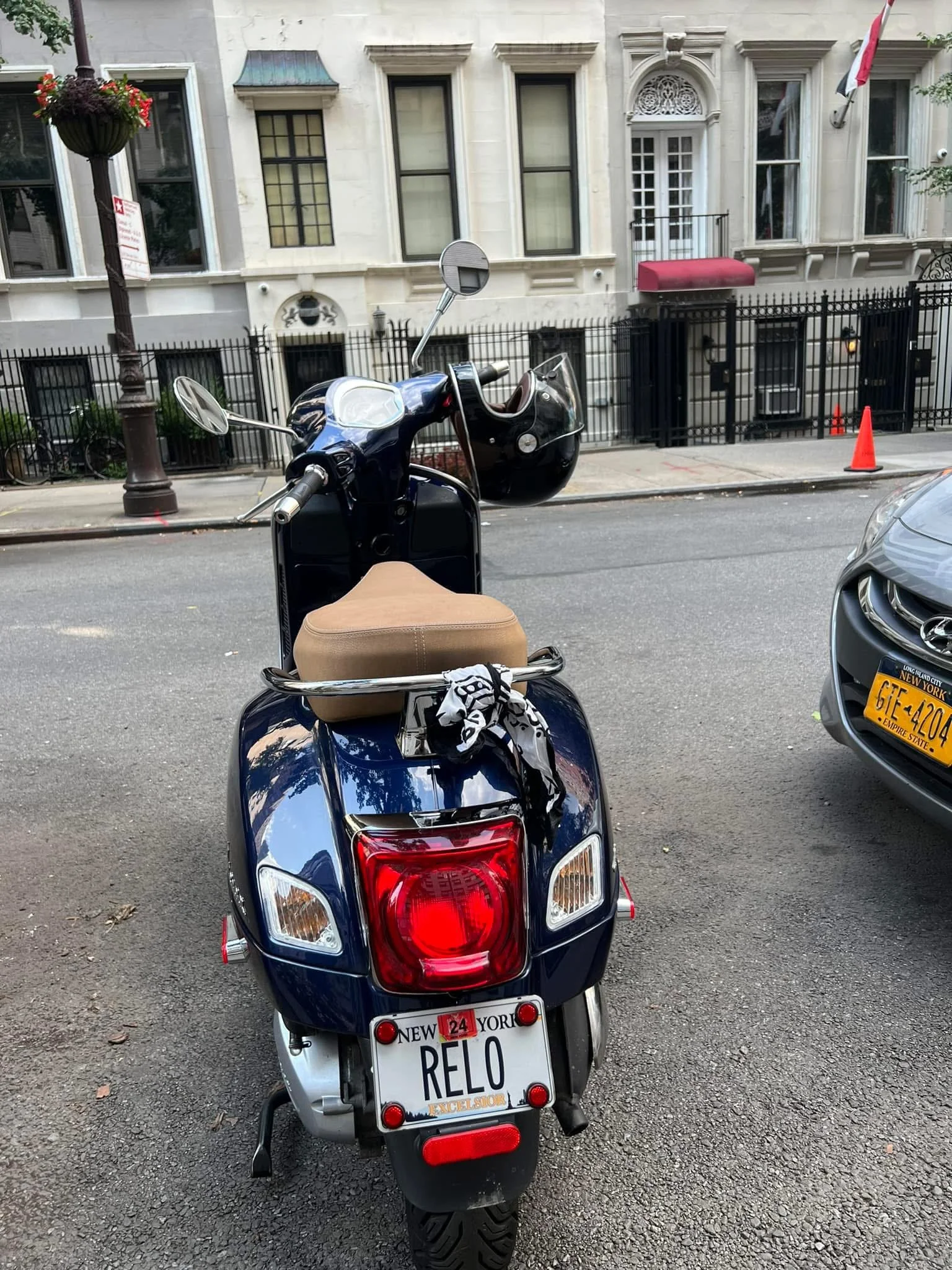 Rear view of a blue scooter with a personalized license plate reading 'RELO' parked on the street. The scooter has a tan seat, a black helmet hanging from the handlebar, and a decorative scarf. Background shows residential buildings with white walls,