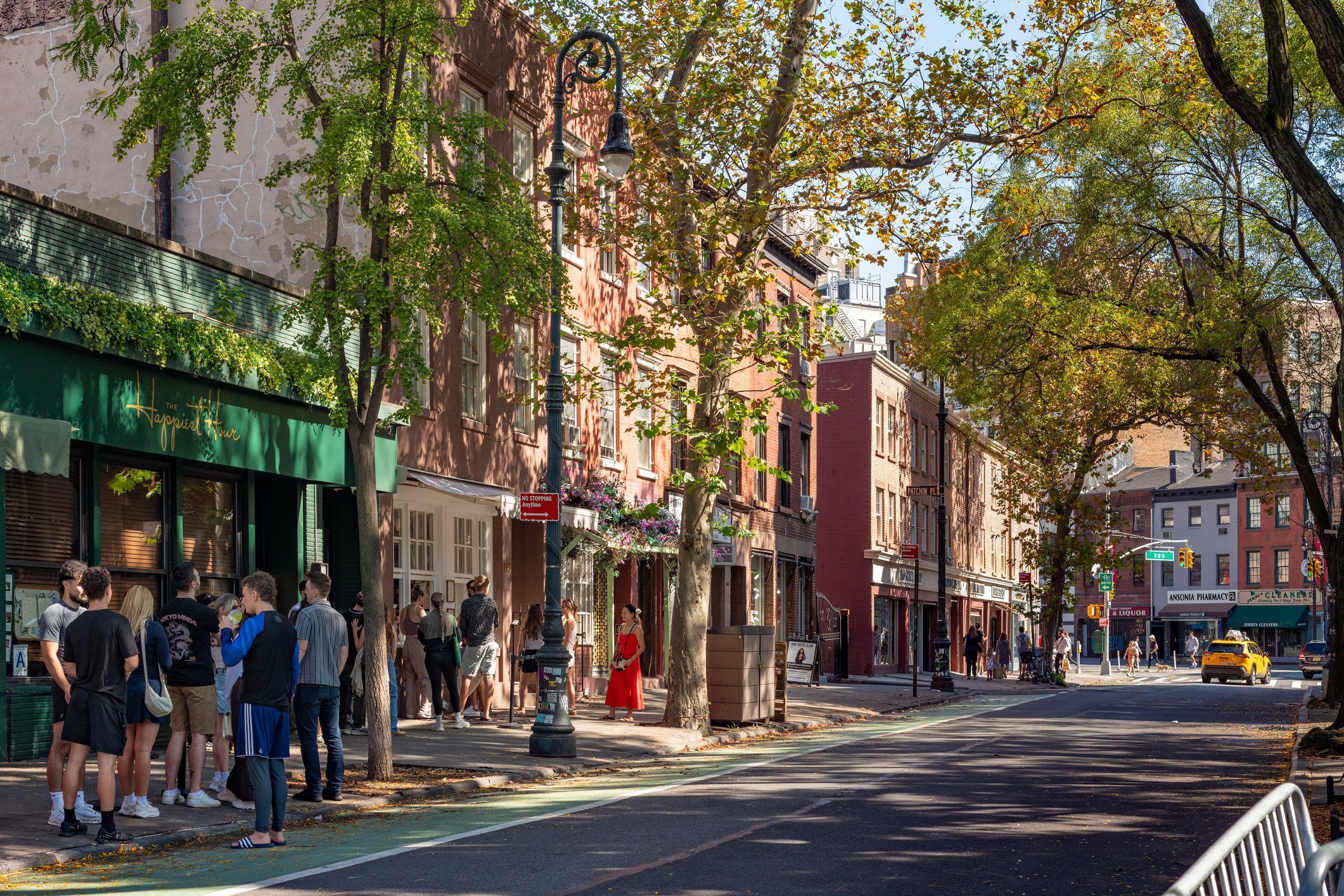 People standing in line outside a building on a city street with trees and colorful buildings.