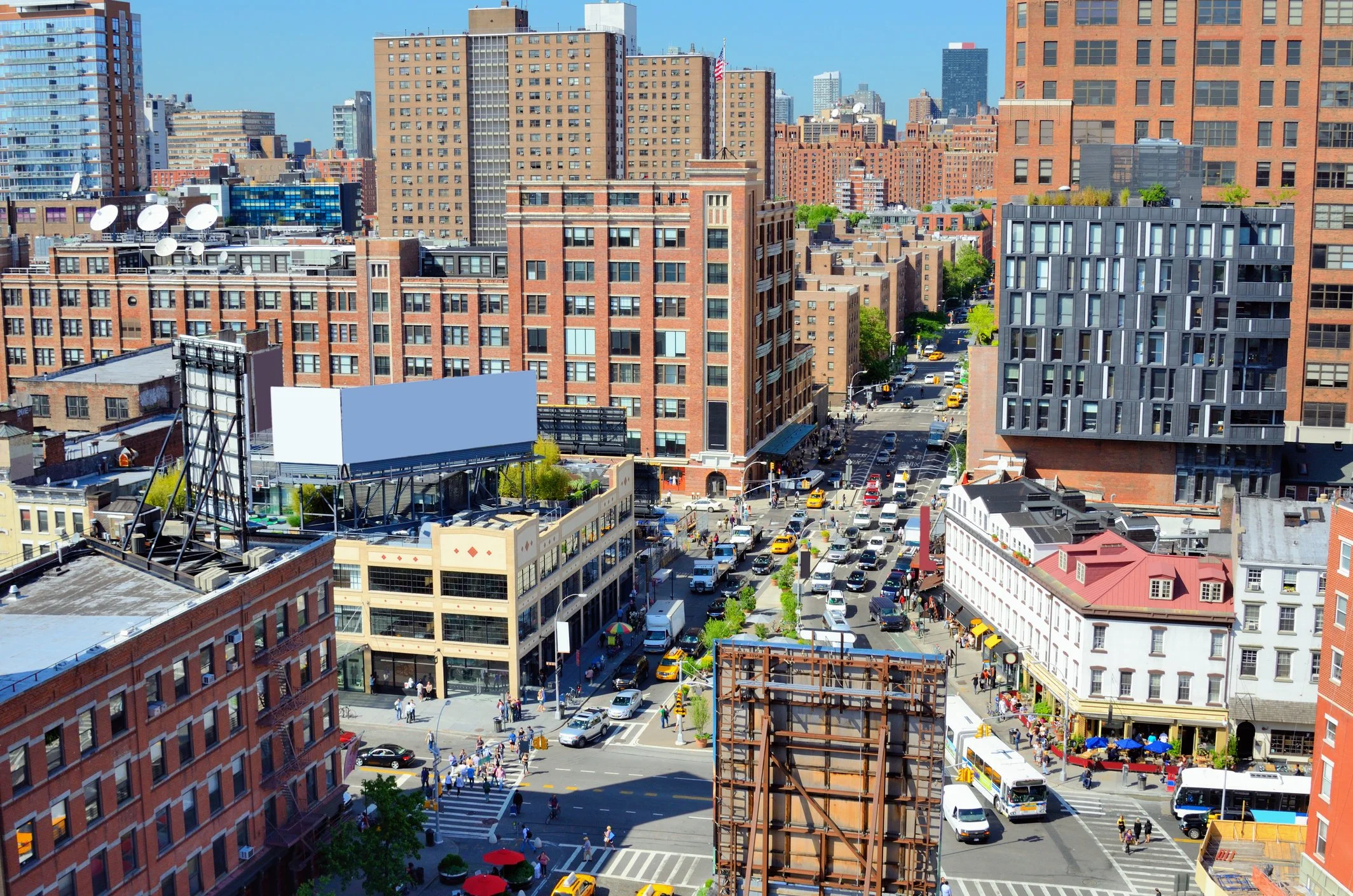 A busy city intersection with numerous red brick and modern office buildings, yellow taxis, and pedestrians on the crosswalks, under a clear blue sky.