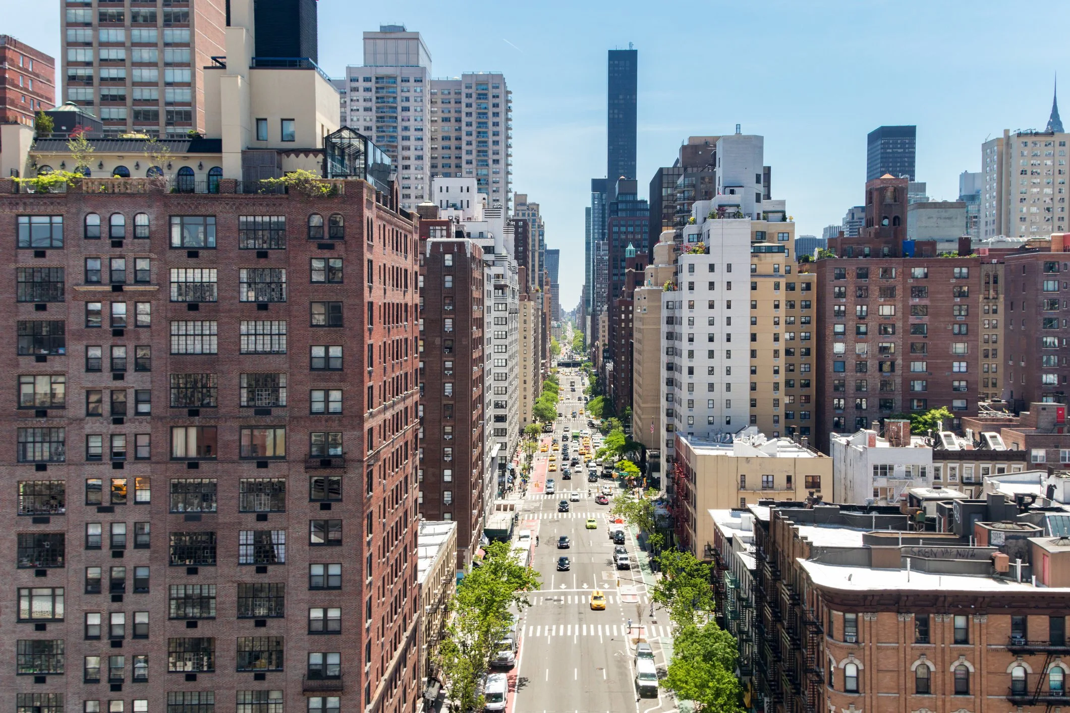 City street lined with tall buildings on both sides, with cars and taxis driving along and trees planted along the sidewalks under a clear blue sky.