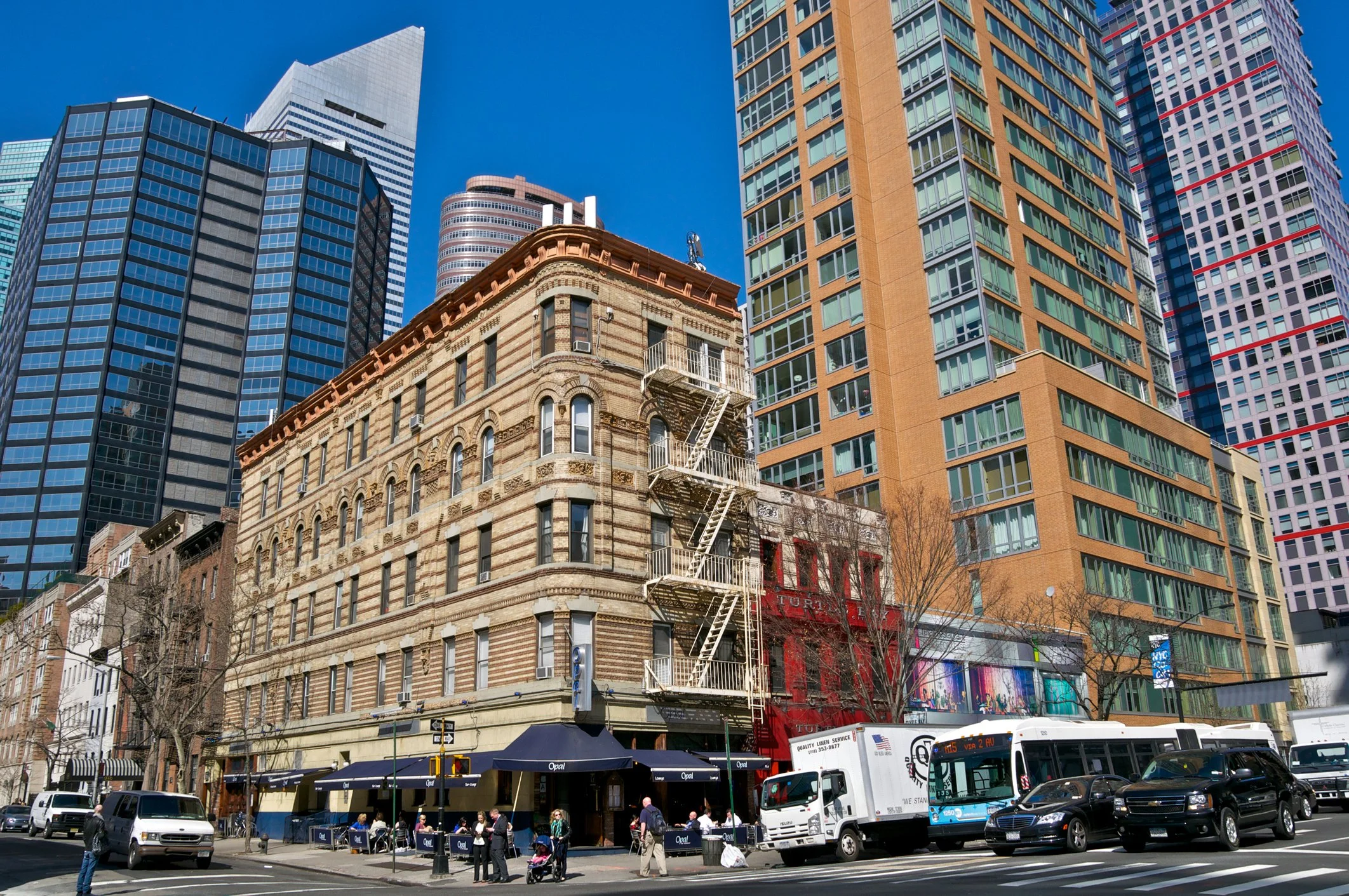 Street scene with crowded sidewalk, tall modern skyscrapers, an older brick building with fire escape, and various parked and moving vehicles.