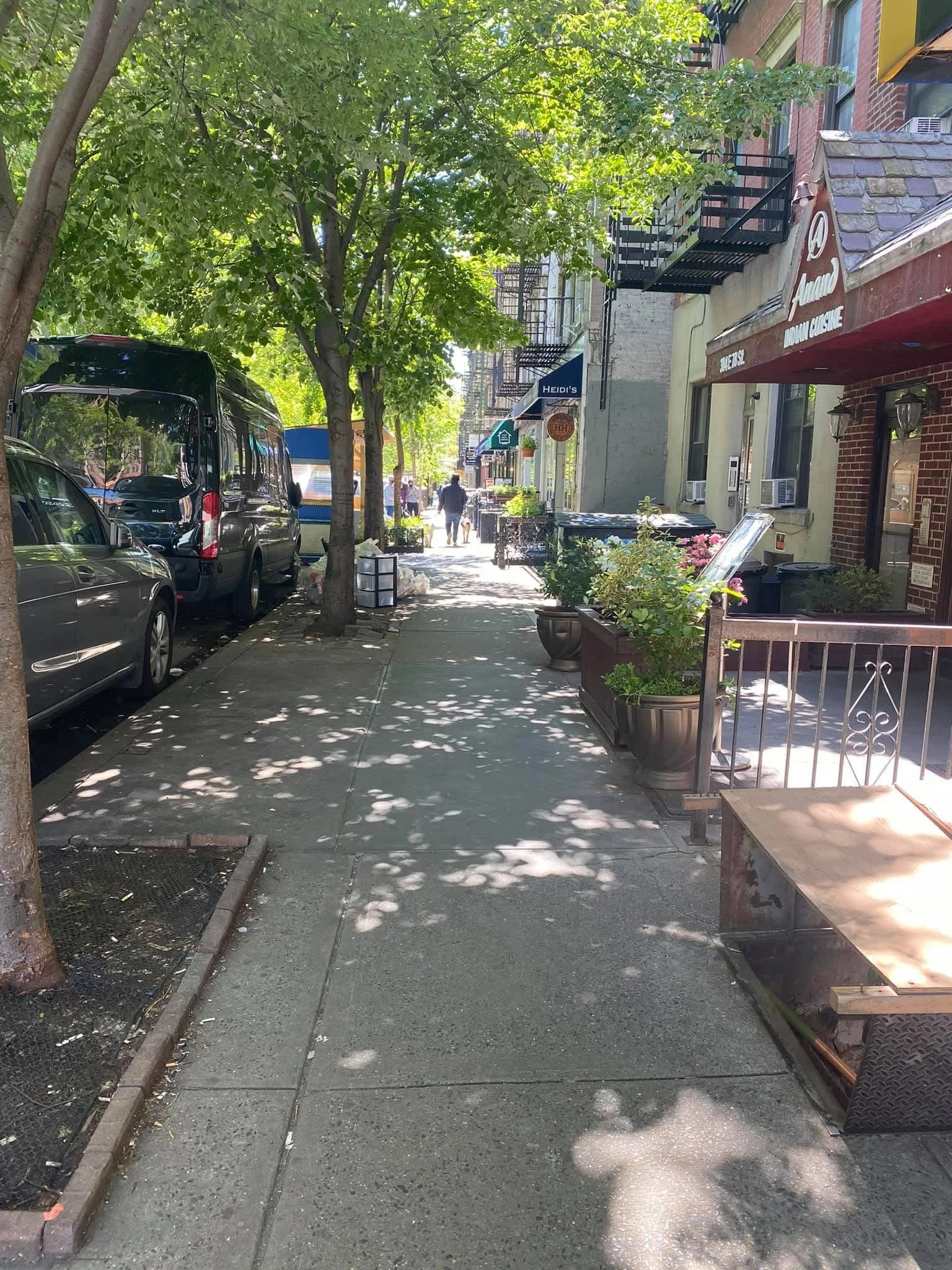 City sidewalk with parked cars, trees casting shadows, storefronts with signs, and pedestrians walking in the distance on a sunny day.