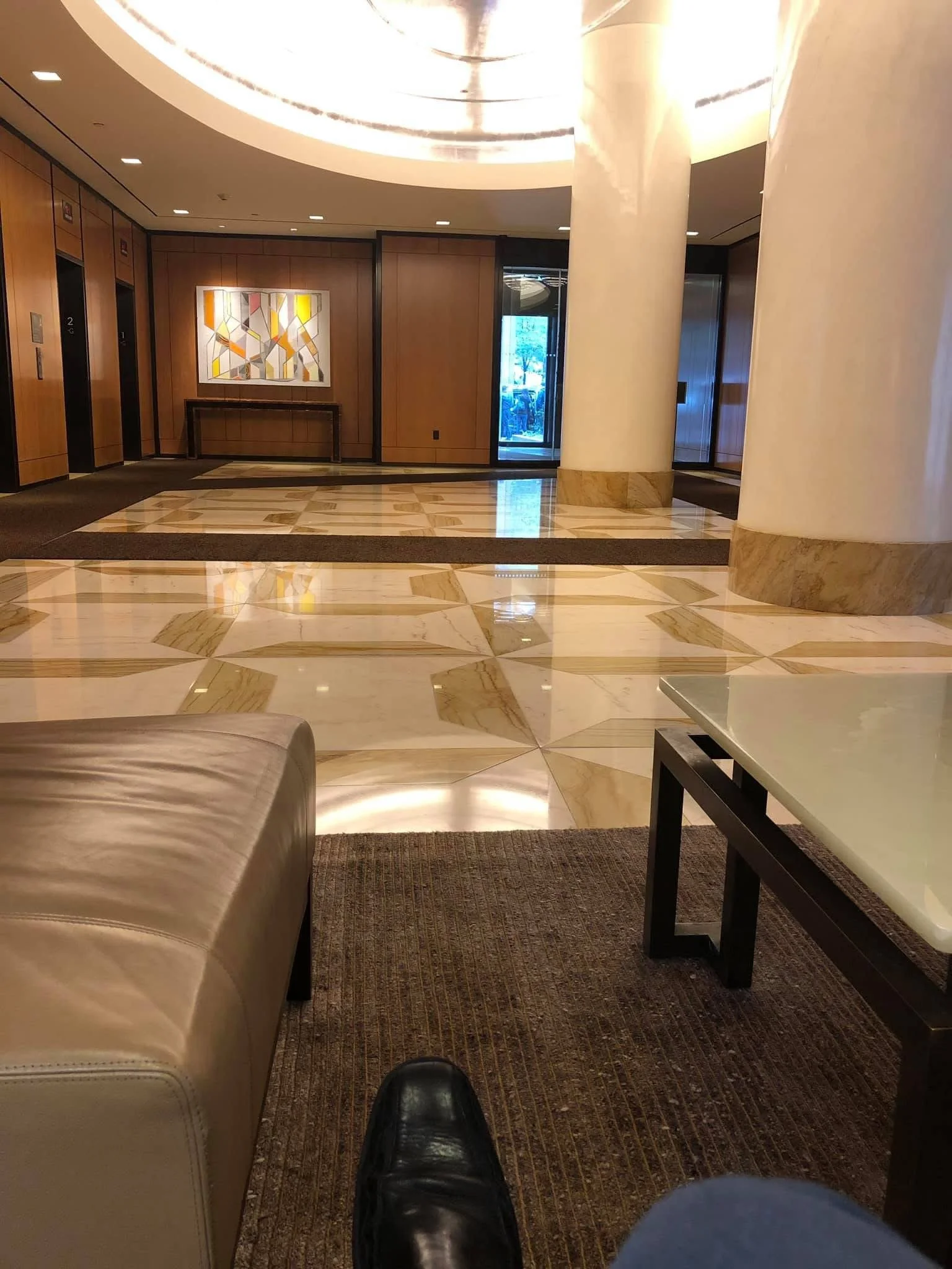Hotel lobby with marble floors, wooden walls, large white columns, elevator doors, artwork on the wall, and a glass entrance door.
