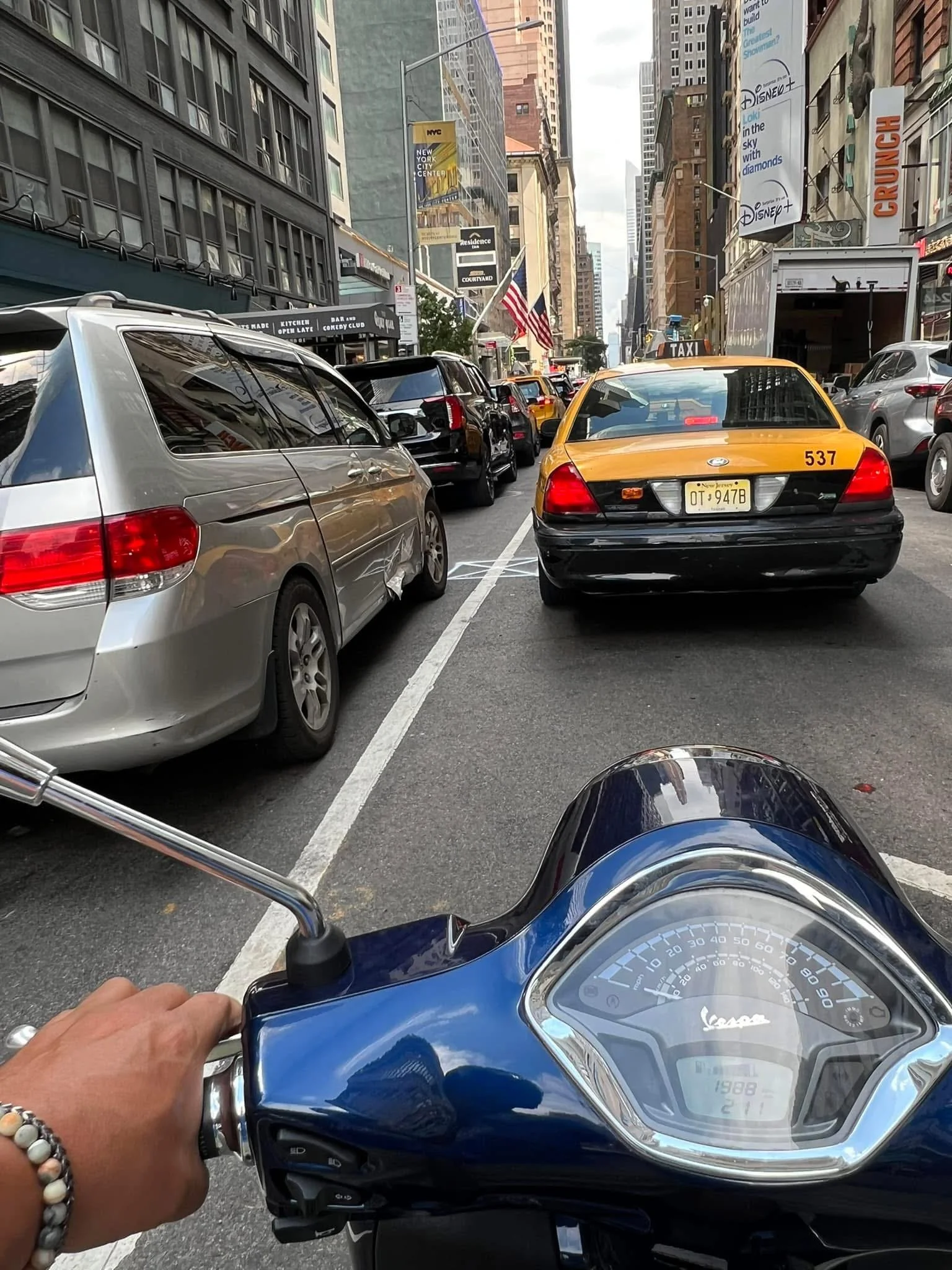 View from a Vespa scooter rider's perspective on a busy city street, showing parked cars, yellow taxis, tall buildings, and storefronts.