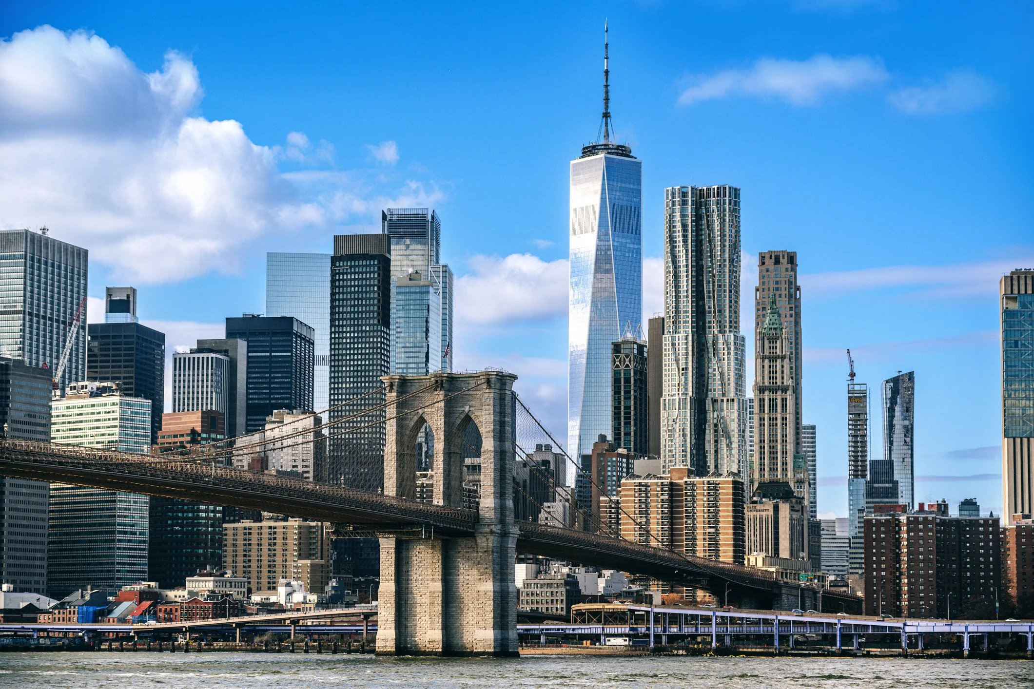 City skyline featuring the Brooklyn Bridge with skyscrapers in New York City, including One World Trade Center, under a partly cloudy blue sky.