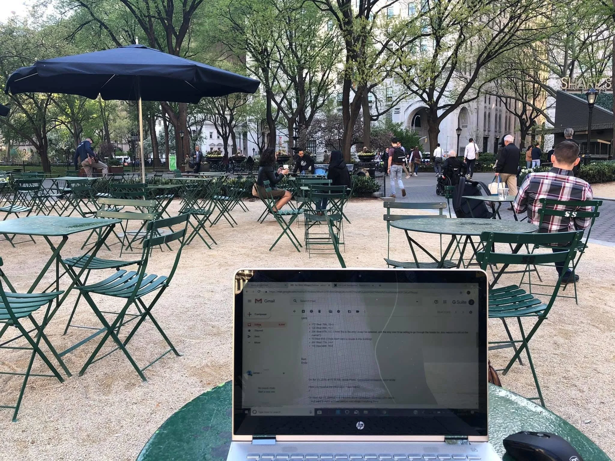 Computer on outdoor table at a park with empty green chairs and tables, people walking and sitting in the background, trees, and buildings.