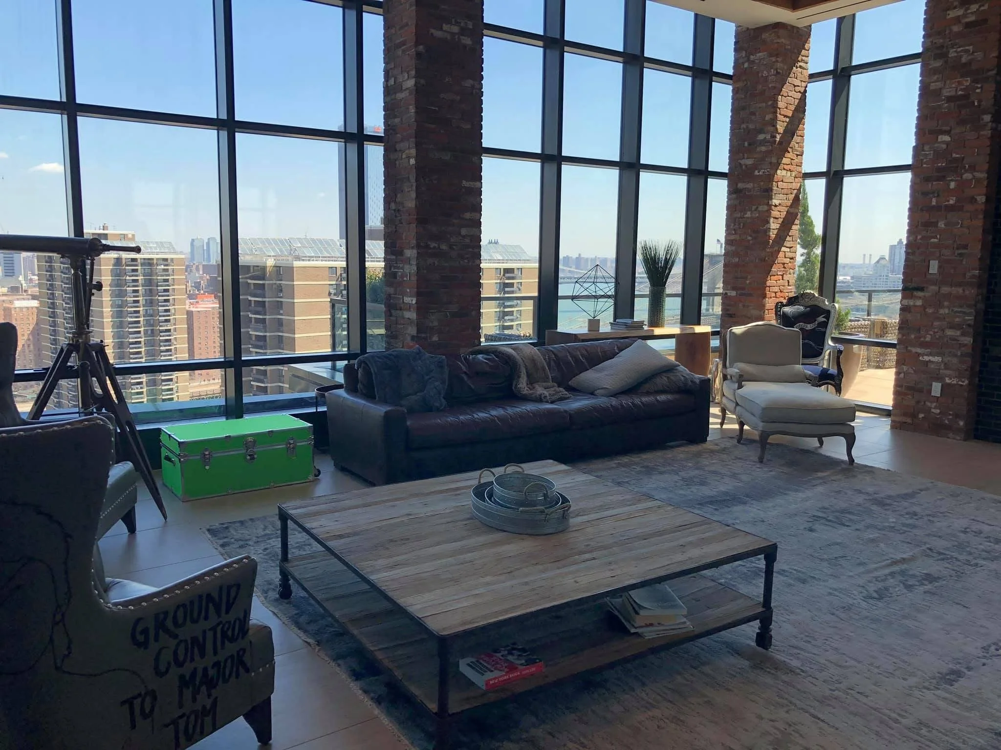 Modern living room with large floor-to-ceiling windows, exposed brick walls, leather sofa, armchair with ottoman, wooden coffee table, telescope, green storage chest, decorative objects, and view of city skyline.
