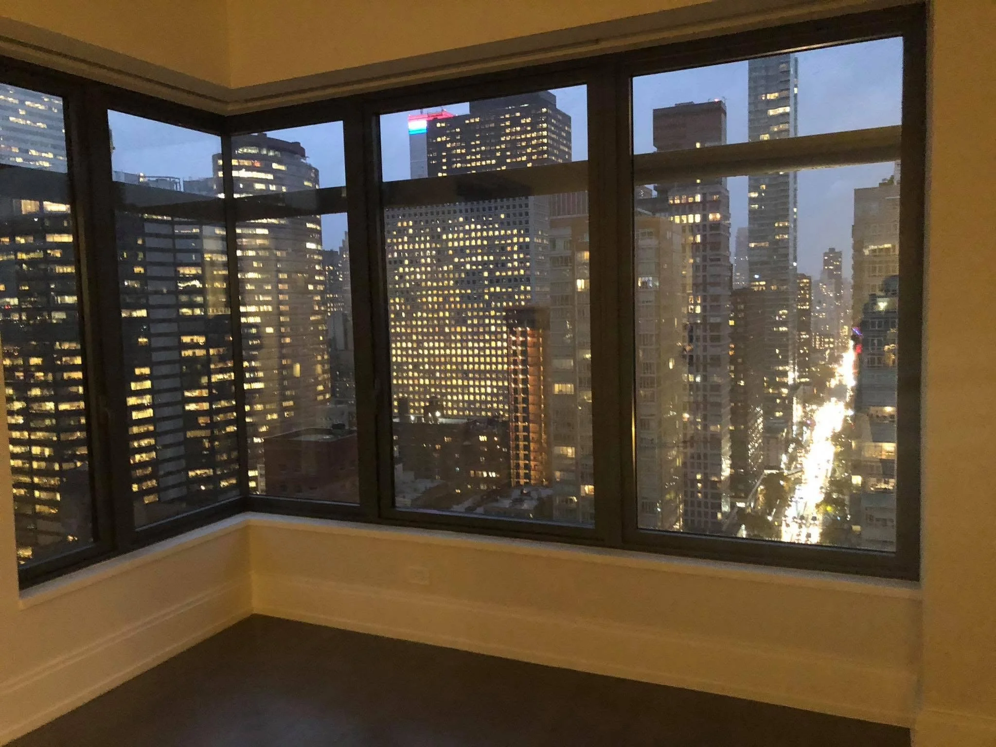 Interior of an empty room with large floor-to-ceiling windows overlooking a city skyline at dusk, with buildings illuminated and a busy street visible.