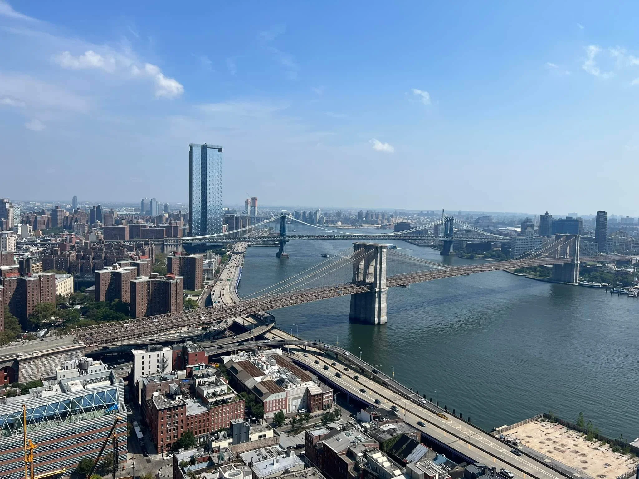 Aerial view of New York City featuring the Brooklyn Bridge crossing the East River with Manhattan skyline in the background under a blue sky with some clouds.