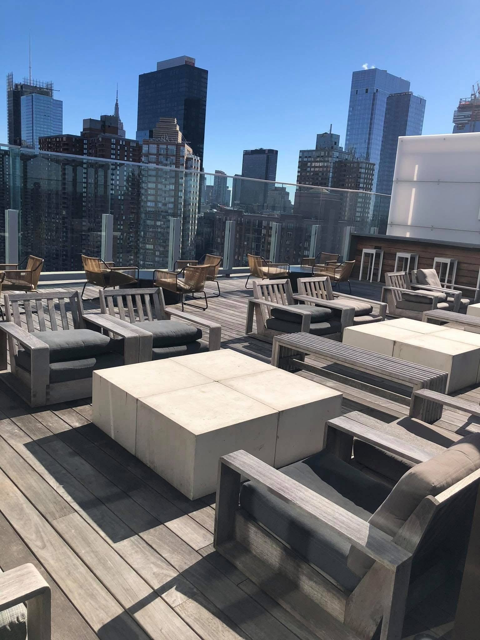 Rooftop patio with wooden furniture, cushioned chairs, and modern decor overlooking a city skyline with tall skyscrapers.