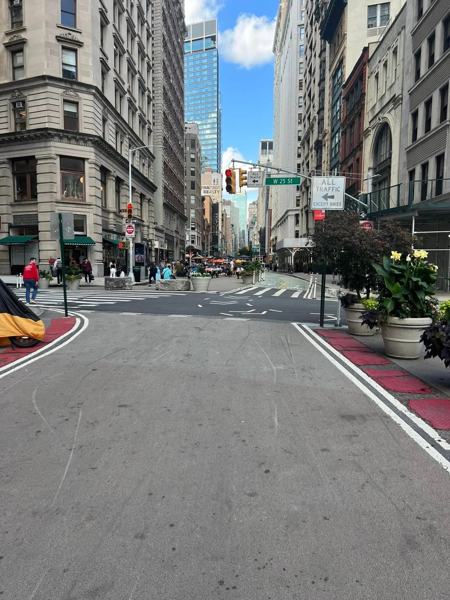 Urban street scene with tall buildings, a crosswalk, and street signs indicating W 25th Street and traffic directions, during daytime with partly cloudy sky.