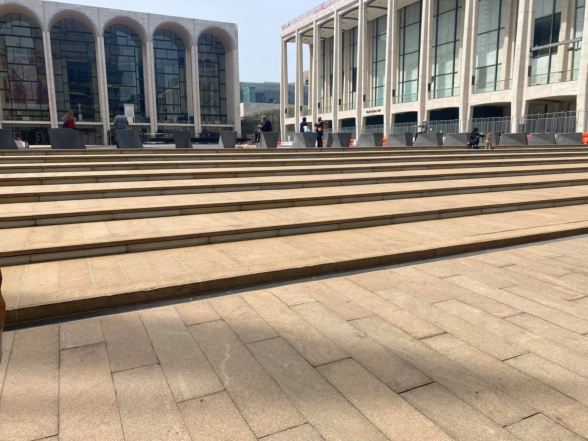 Exterior view of a public space with wide steps leading up to a modern building with large glass windows and arched architecture, with a few people walking or sitting around.