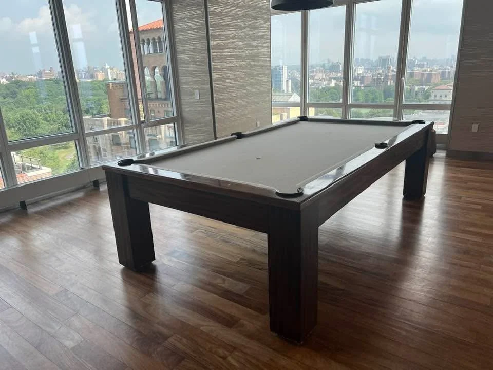 A sunlit indoor room with a pool table placed on a hardwood floor, large windows showing cityscape and greenery outside.