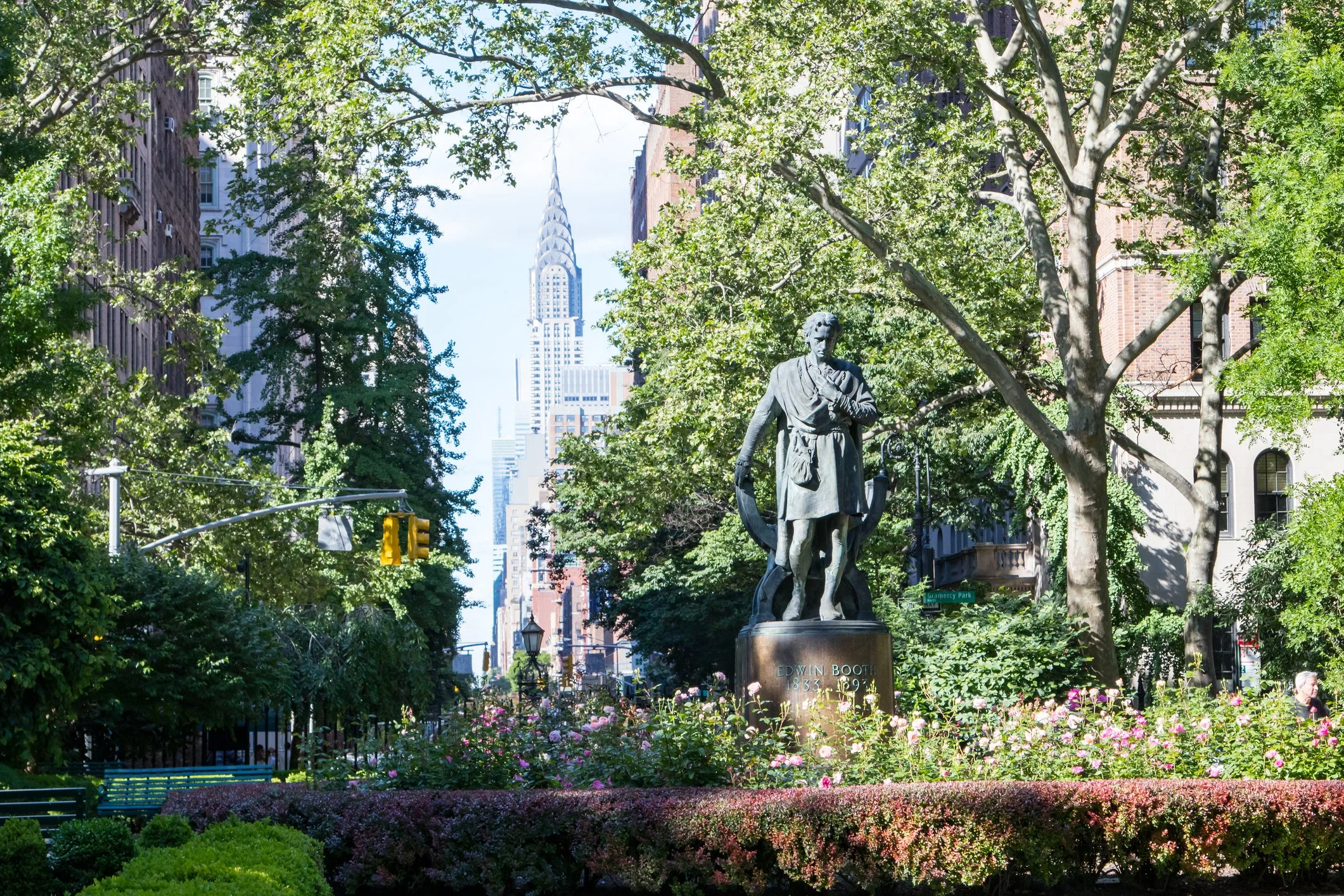 Statue of Edwin Booth in a park surrounded by flowers and trees, with tall buildings including the Chrysler Building in the background, in New York City.